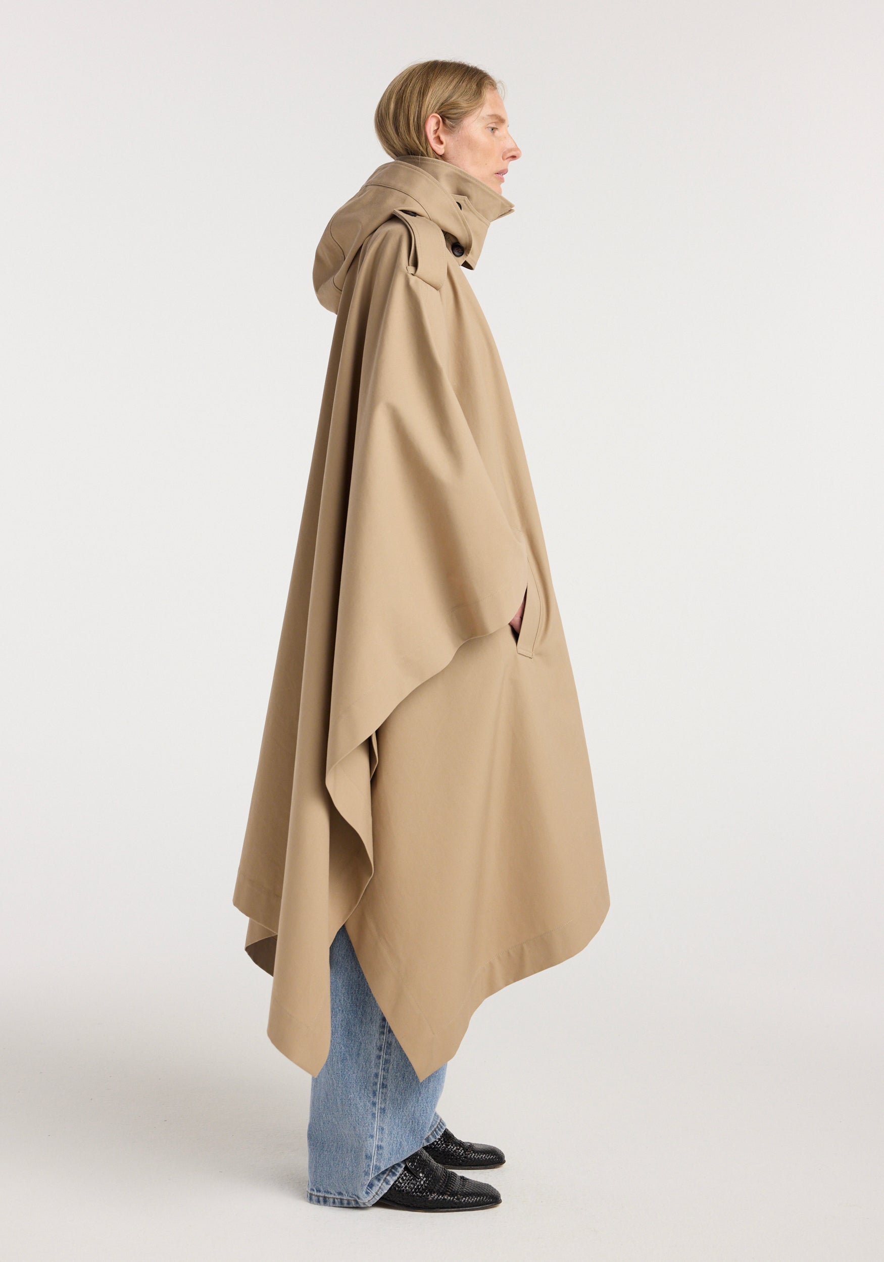 Side view of a person wearing a beige hooded cape trenchcoat | beige with long, flowing fabric over light blue jeans and black shoes against a plain background.
