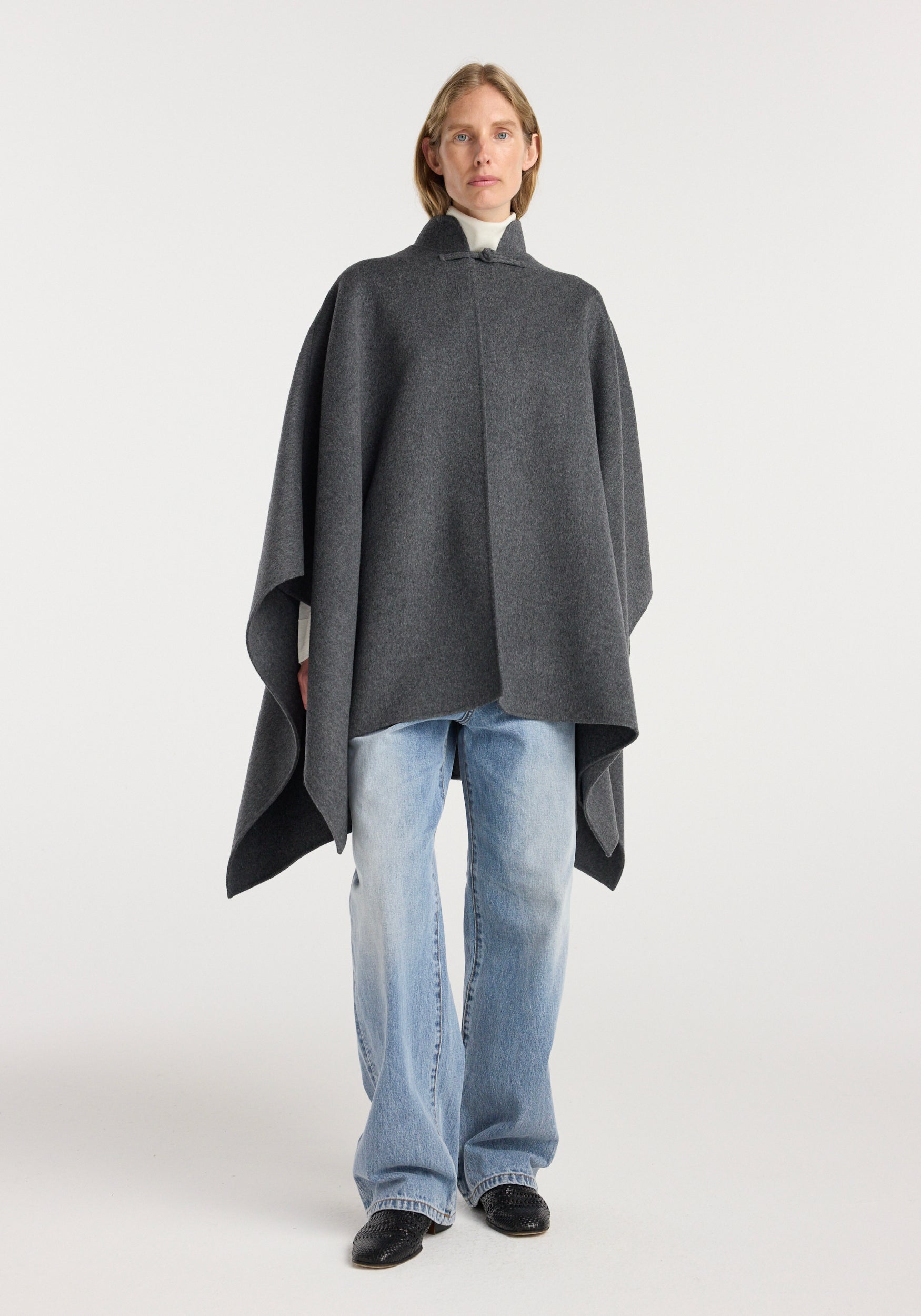 Model wearing a Double-faced wool pankou closure poncho | dark grey melange over a white turtleneck paired with light blue jeans and black shoes, standing against a plain background.