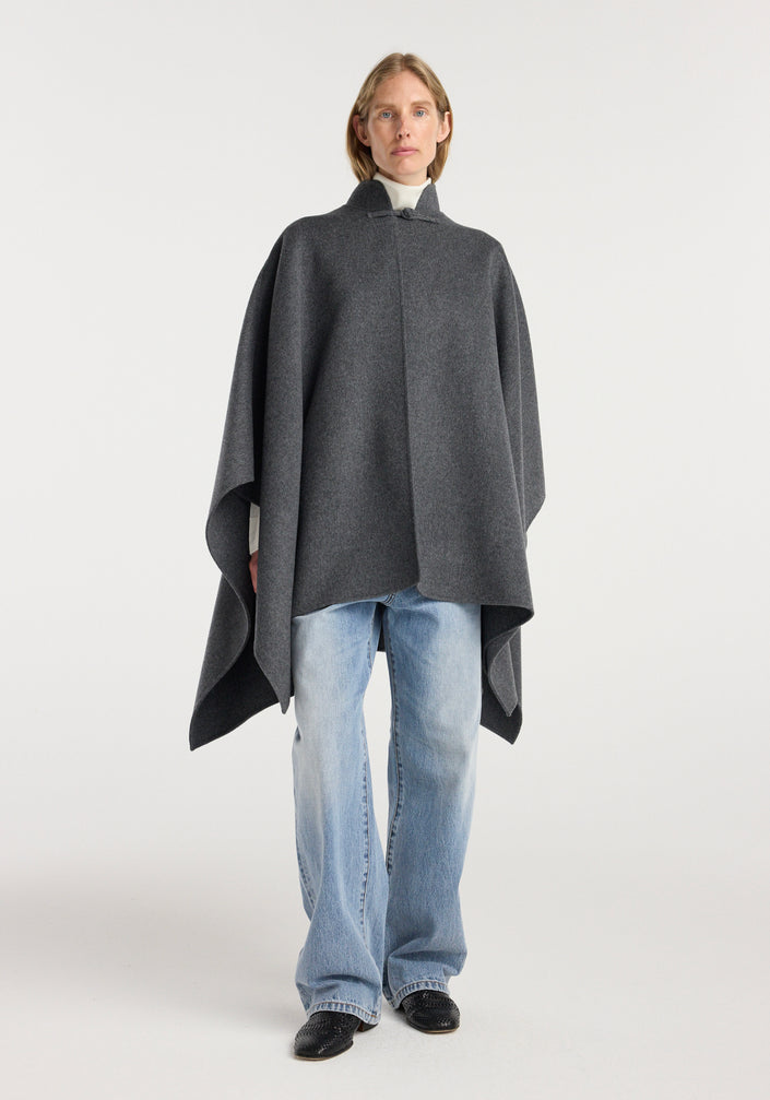 Model wearing a Double-faced wool pankou closure poncho | dark grey melange over a white turtleneck paired with light blue jeans and black shoes, standing against a plain background.