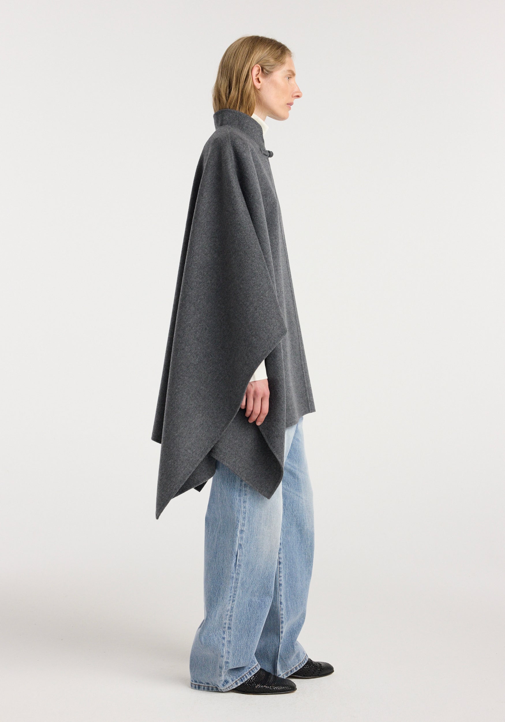 Side profile of a person wearing a double-faced wool pankou closure poncho | dark grey melange over a white top with light blue jeans and black shoes, standing against a plain background.