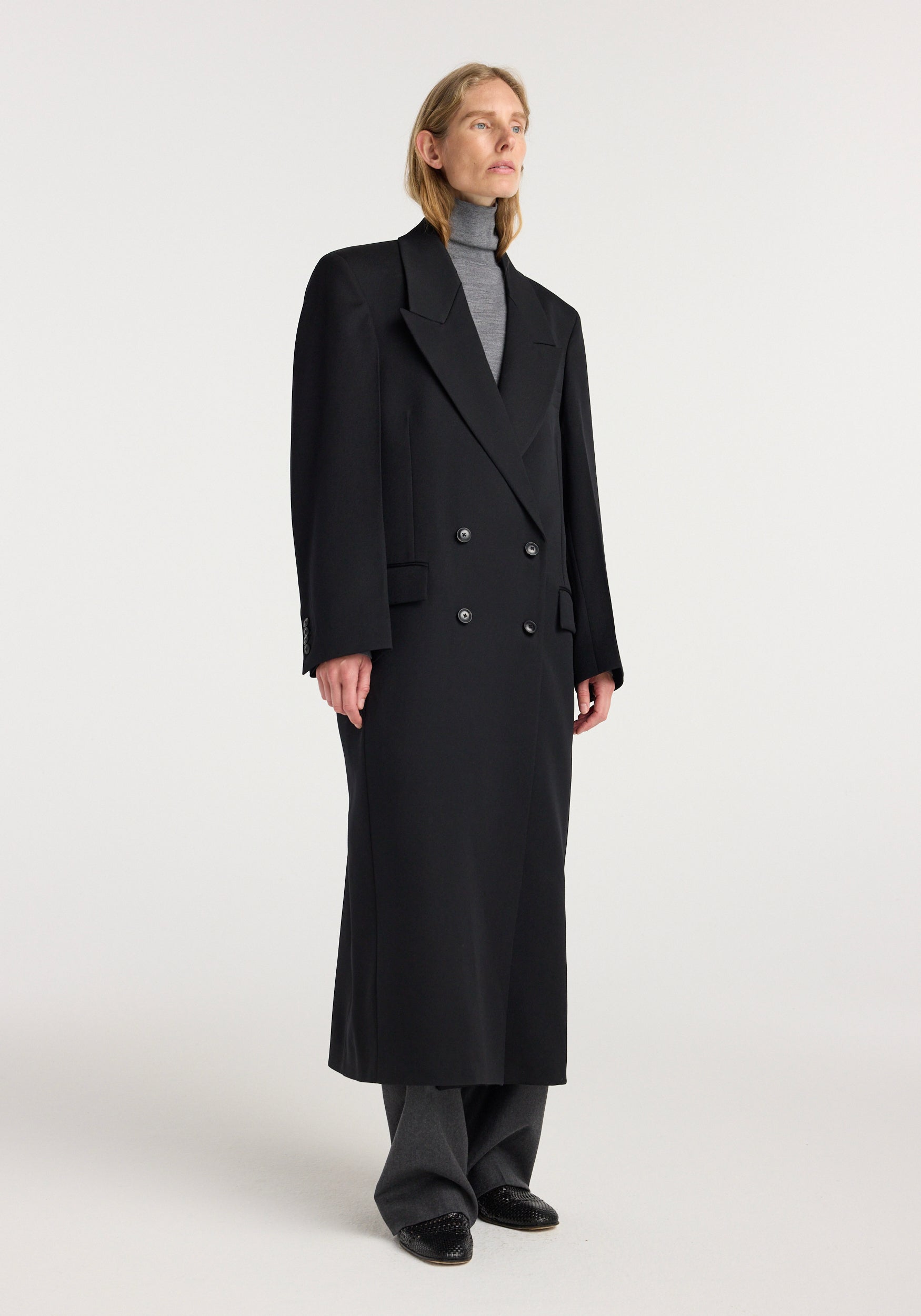 Woman wearing a long black double breasted tailored coat | black over a gray turtleneck and wide-leg pants, standing against a plain light background.