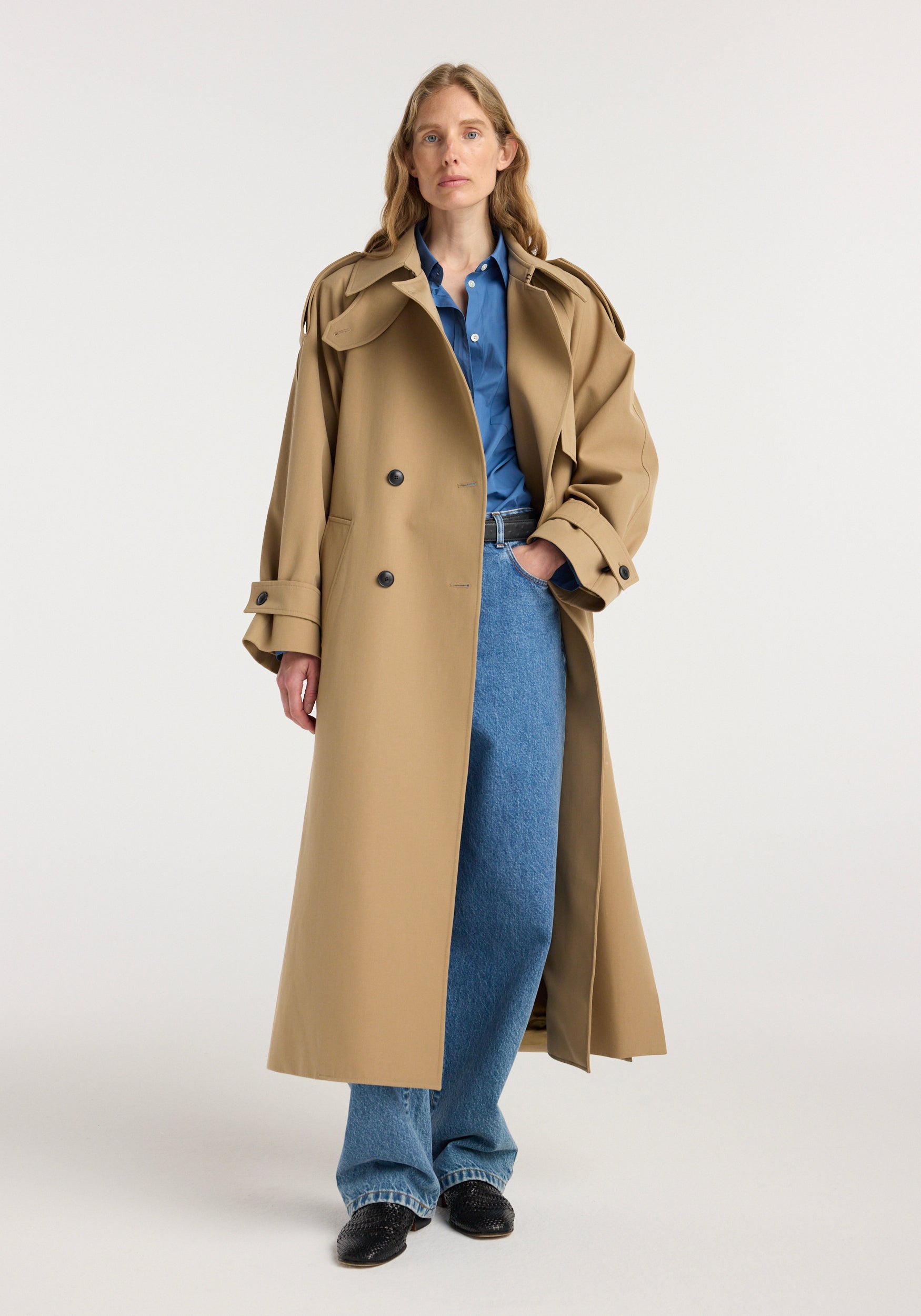 Woman wearing a beige Classic trench | beige over a blue shirt and jeans, standing against a plain white background with hands in pockets.
