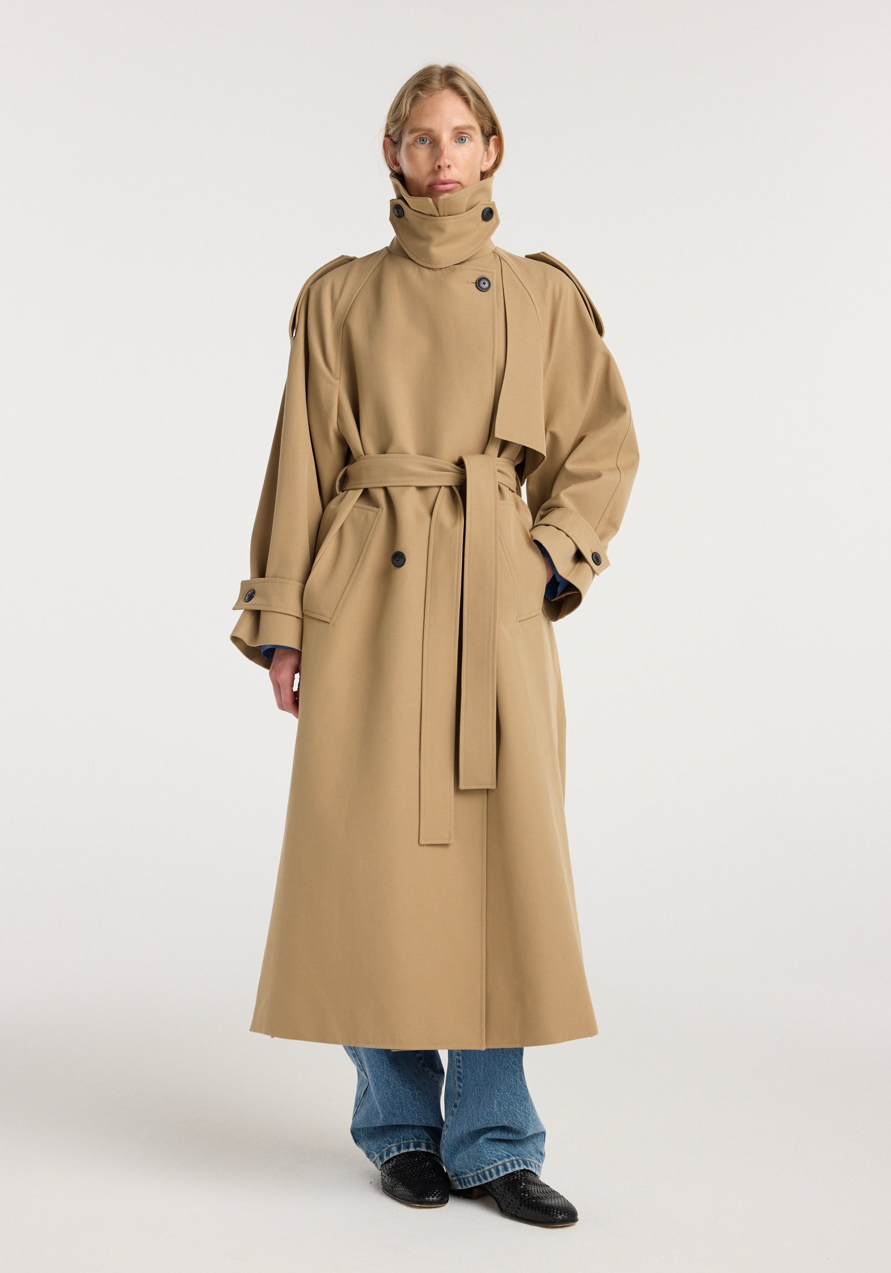 Woman wearing a beige Classic trench | beige with a high collar, belted waist, and button details, styled with blue jeans and black shoes against a plain background.