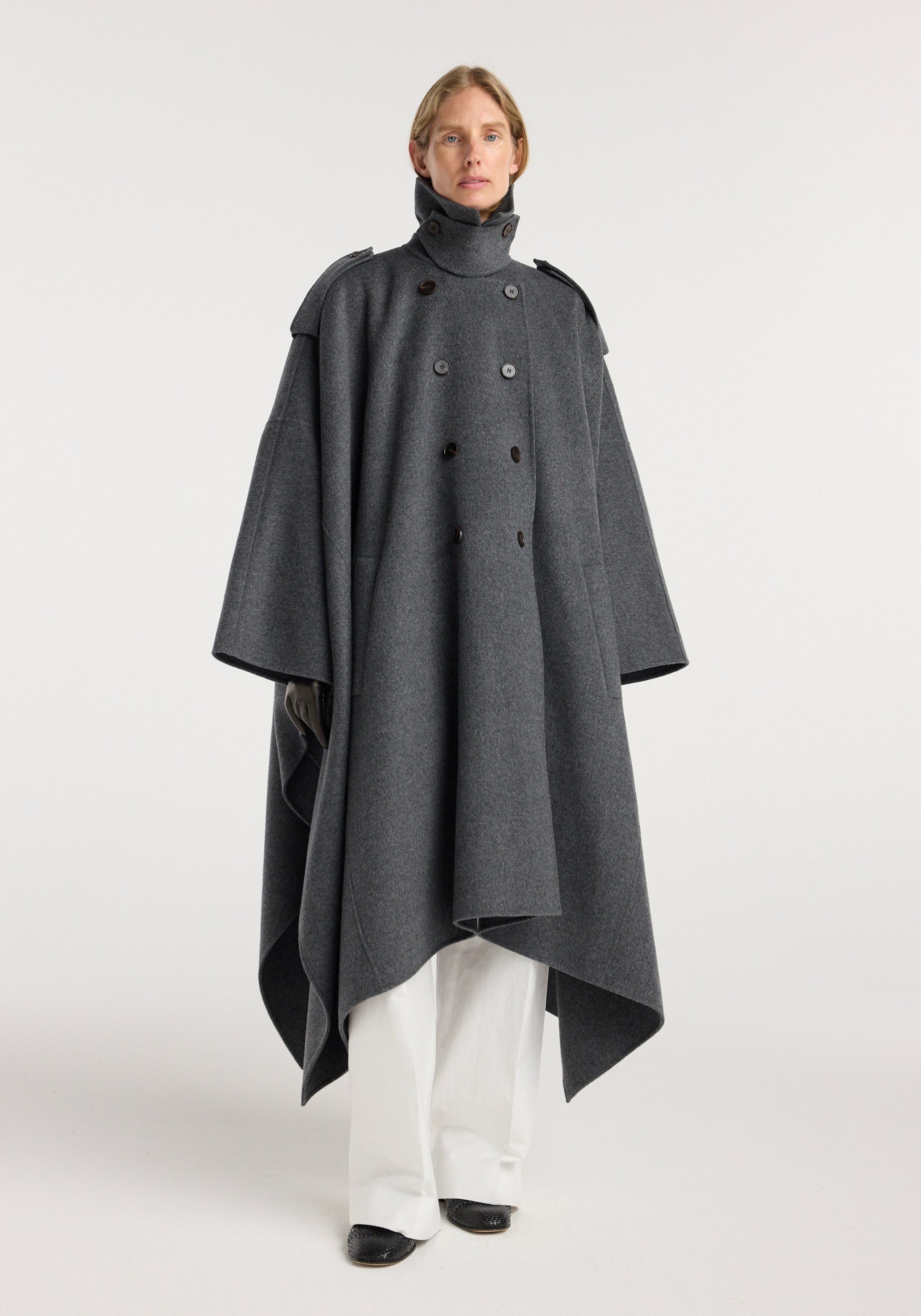 A model wearing a double-faced cape trenchcoat | dark grey melange, featuring a high neck, shoulder epaulettes, double-breasted button closure, wide draped sleeves, and an asymmetrical hem over white pants and black shoes.
