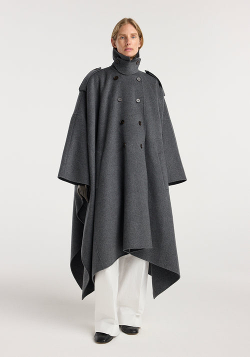 A model wearing a double-faced cape trenchcoat | dark grey melange, featuring a high neck, shoulder epaulettes, double-breasted button closure, wide draped sleeves, and an asymmetrical hem over white pants and black shoes.