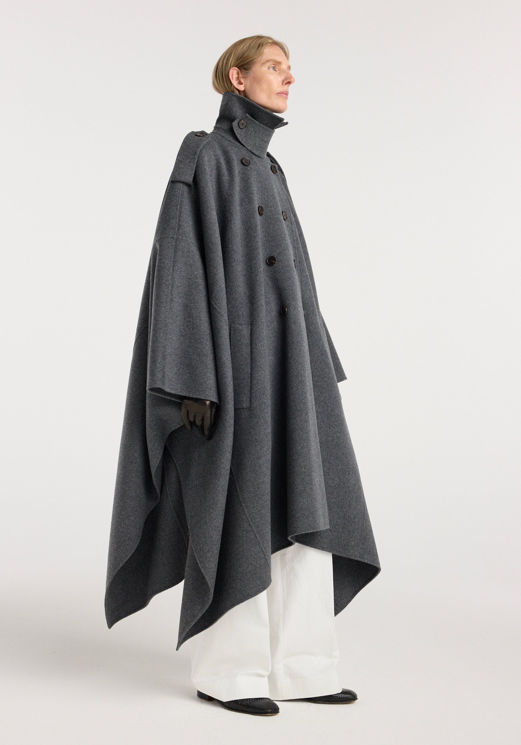Woman wearing a high-collared, dark grey melange double-faced cape trenchcoat with button details, styled with white wide-leg pants and black gloves, standing sideways.