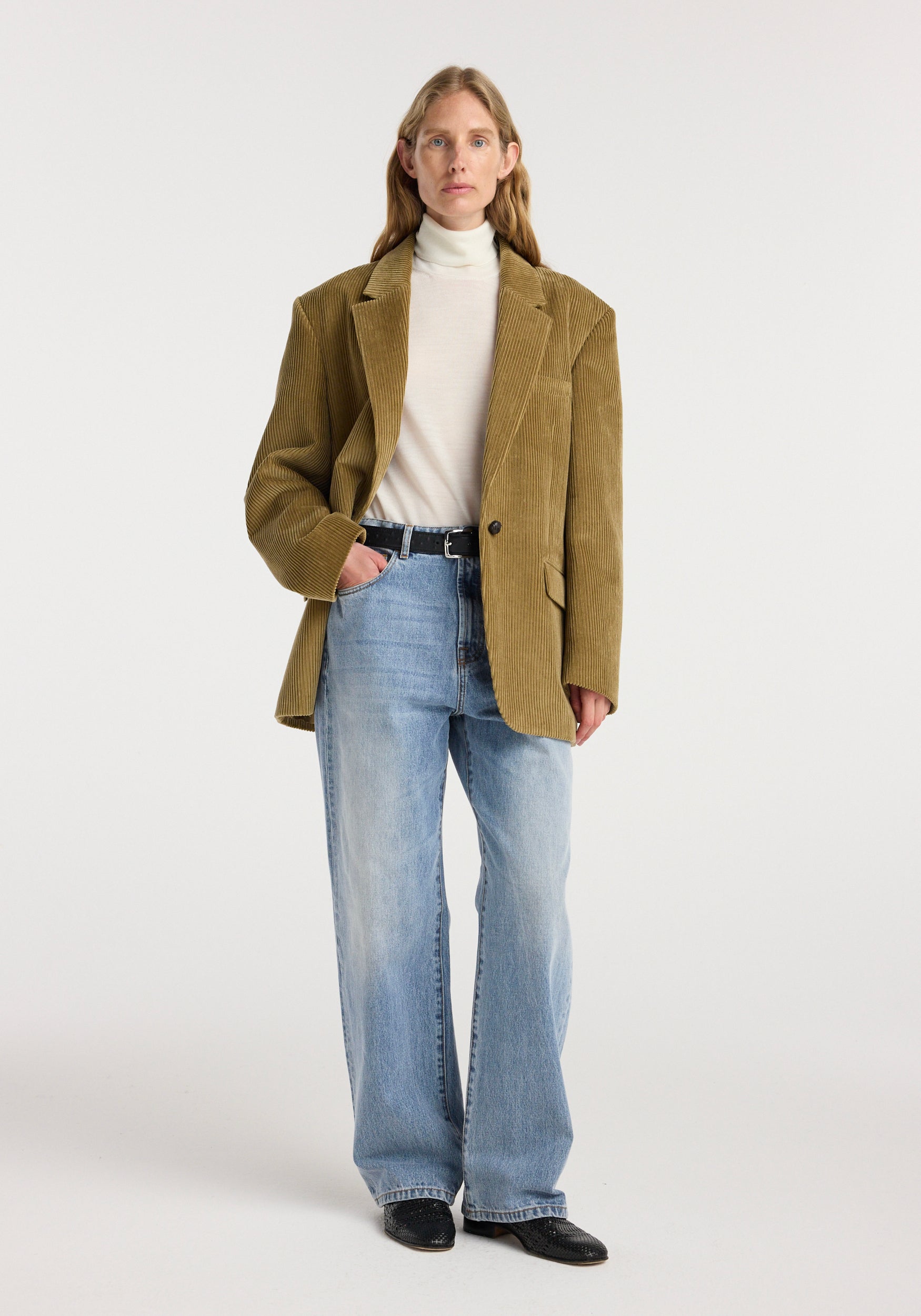 Woman wearing an Oversized corduroy blazer jacket | beige paired with a white turtleneck, light blue wide-leg jeans, and black shoes, standing against a plain white background.