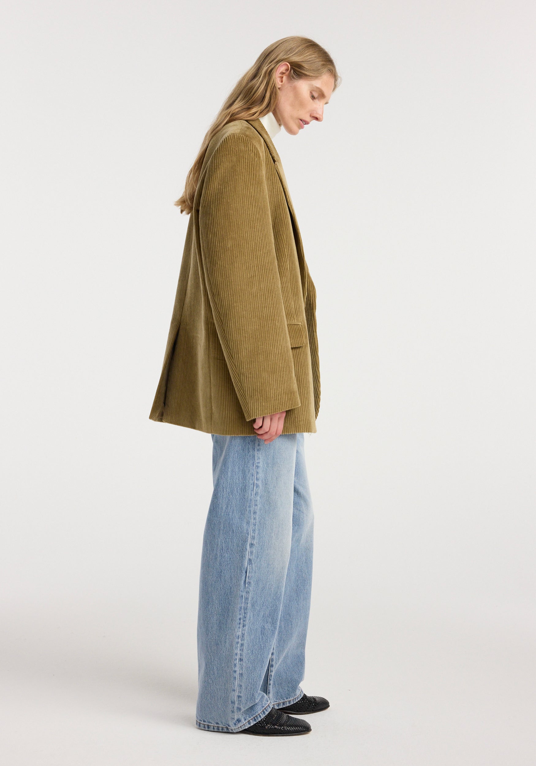 Side view of a woman wearing an oversized corduroy blazer jacket | beige paired with light blue wide-leg jeans and black shoes, standing against a plain white background.