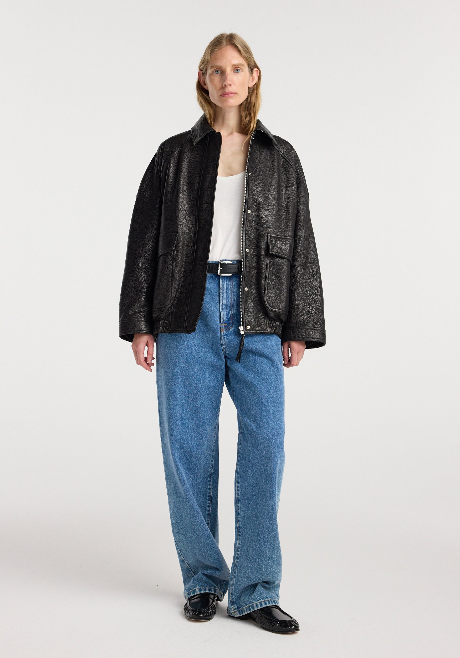 Woman wearing a black grain leather aviator jacket | black over a white top and blue jeans, standing against a plain light background.