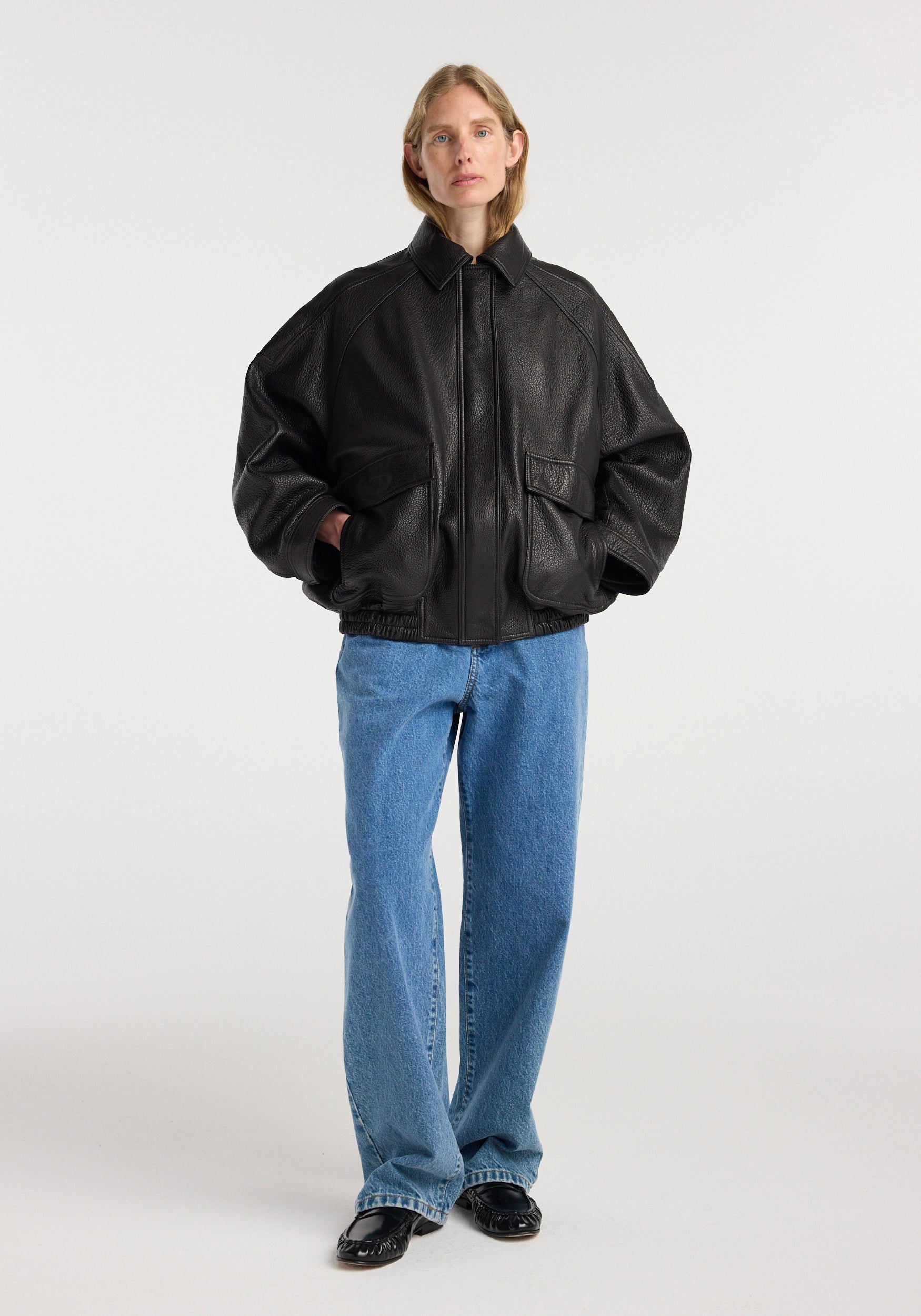 Woman wearing a black grain leather aviator jacket | black with large front pockets and relaxed fit paired with blue jeans and black shoes against a white background.