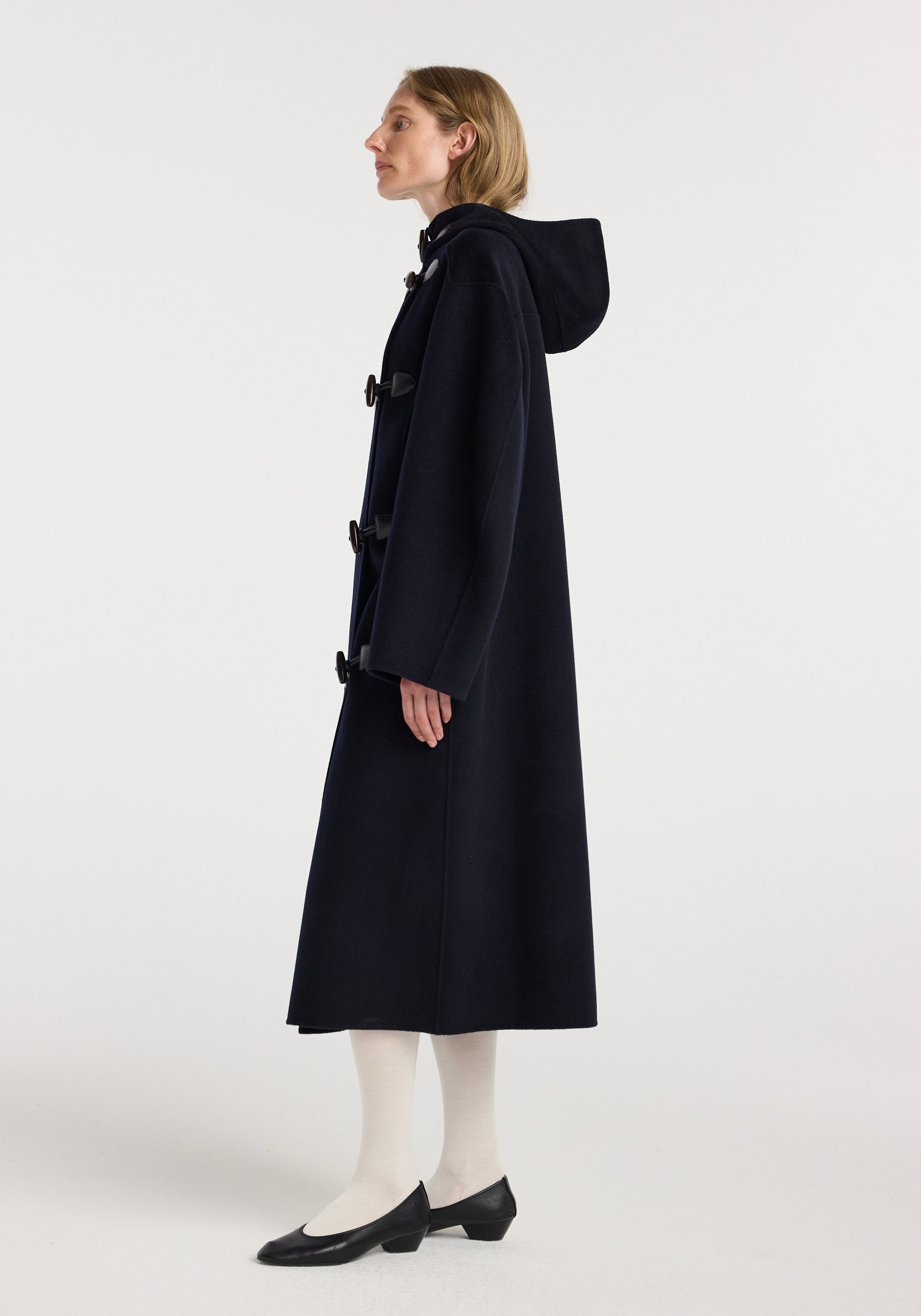 Side view of a woman wearing a long navy coat with toggle fastenings and a hood, styled with white tights and black low-heeled shoes, featuring the Double-faced wool duffel coat | navy.