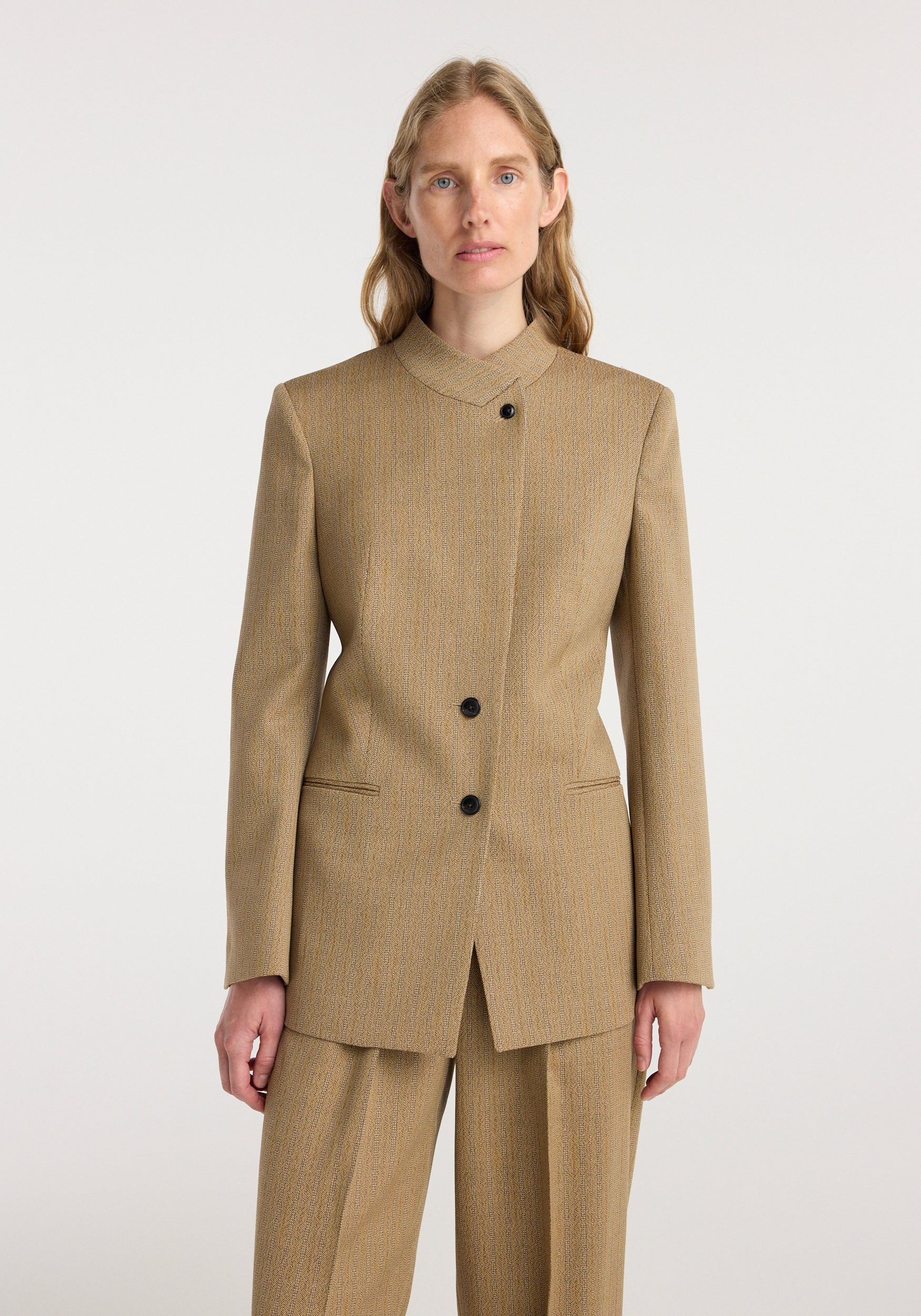 Woman wearing a tailored Overlap lapel blazer | heritage stripe in beige with subtle stripes, featuring an asymmetrical button closure and matching trousers.