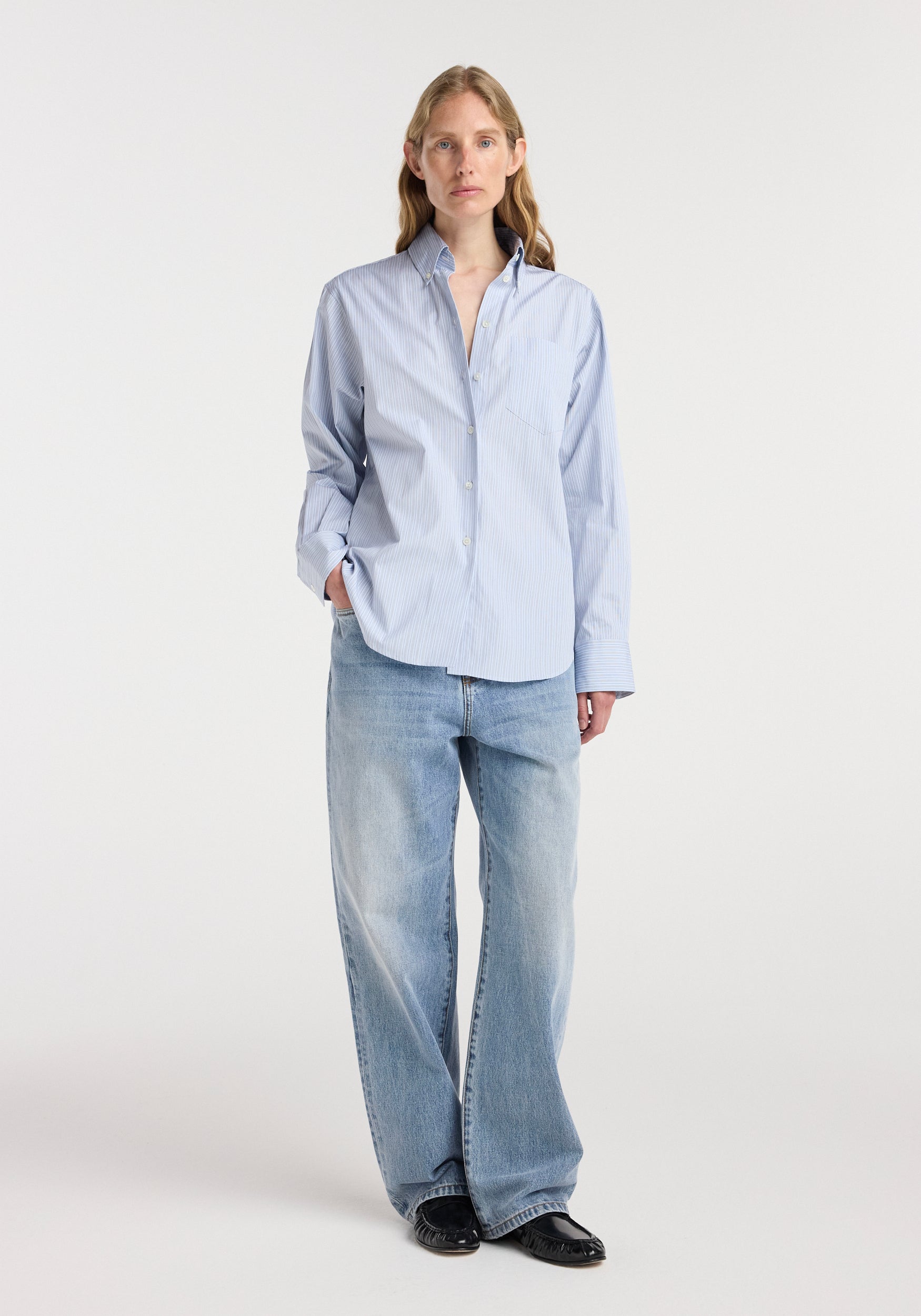 Woman wearing a light blue Oxford striped shirt | blue stripe with a classic collar and button front, paired with loose-fitting light wash jeans and black shoes, standing against a plain background.