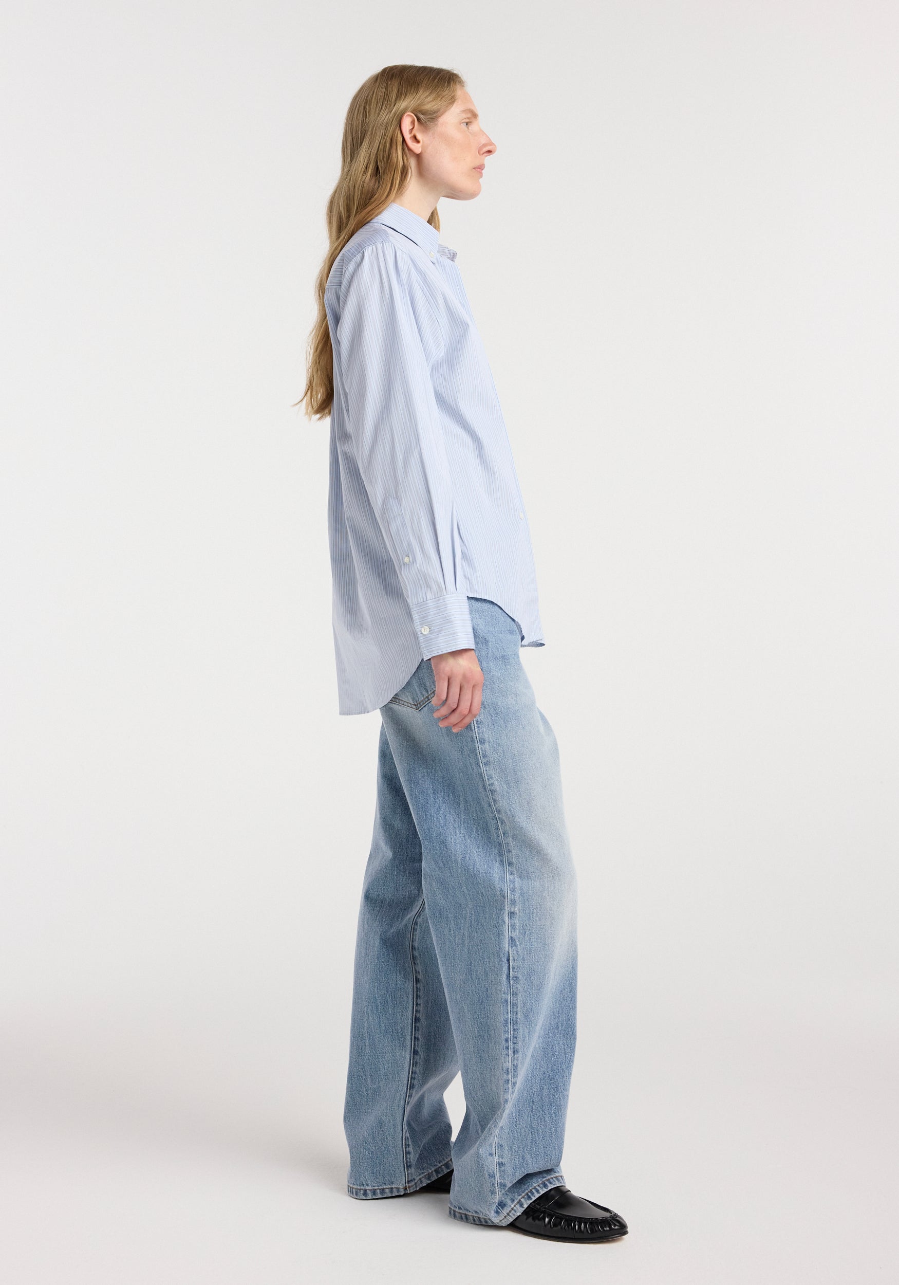 Woman wearing a light blue Oxford striped shirt | blue stripe paired with loose-fitting light wash jeans and black shoes, standing in profile against a plain background.