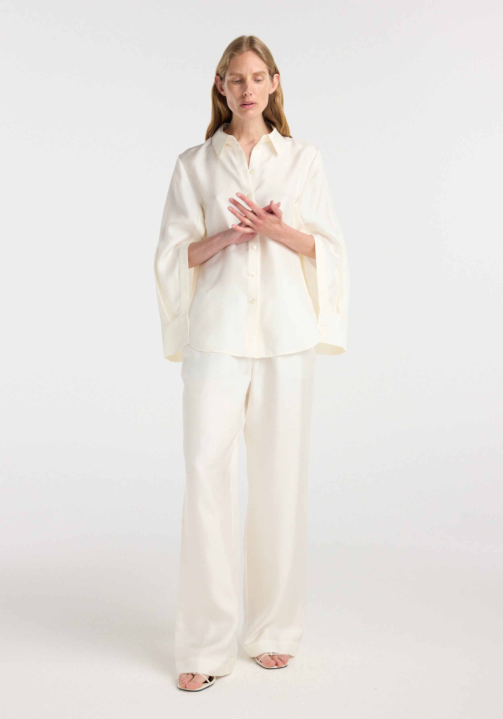 Woman wearing a cream silk open-sleeve detailed shirt | cream with long, flowing sleeves and matching loose pants, standing against a plain white background.