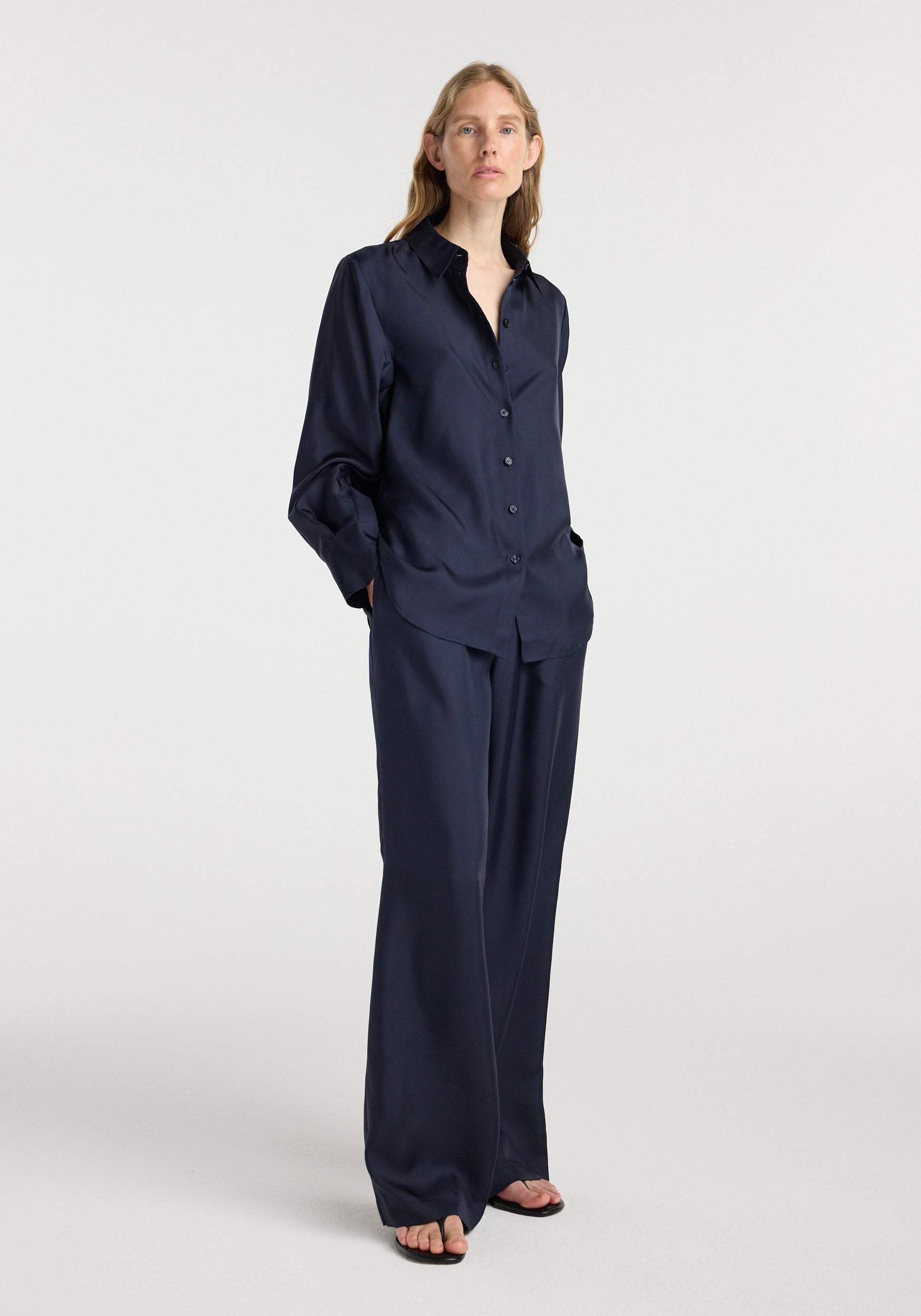 Woman wearing a dark navy silk open-sleeve detailed shirt | night with matching wide-leg pants, standing with hands in pockets against a plain light background.