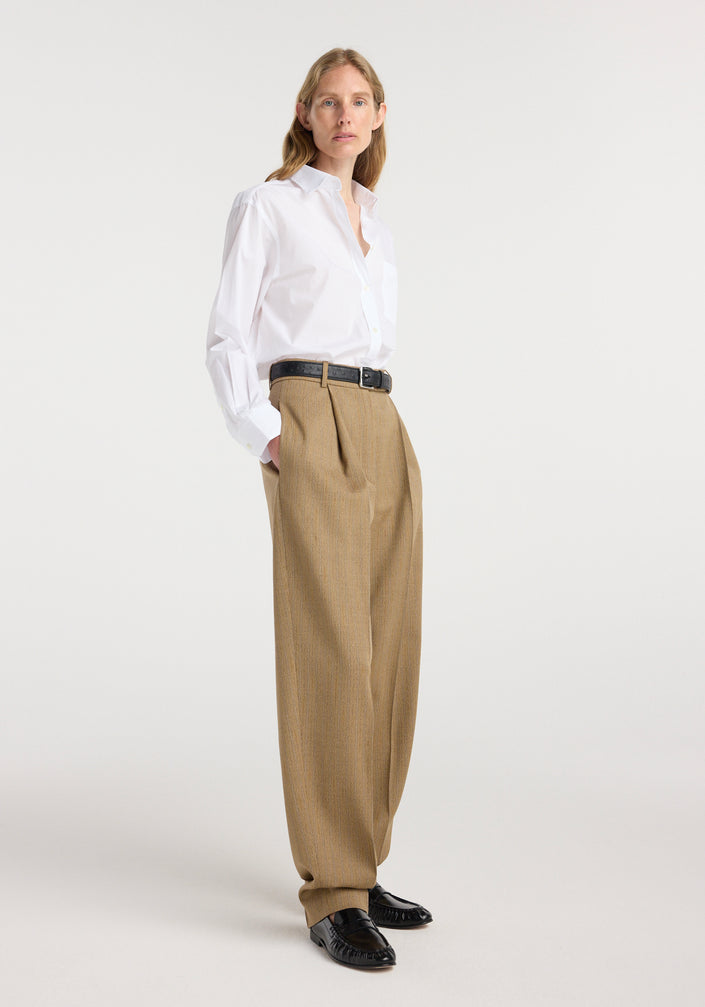 Woman wearing a Classic fit shirt | optic white with long sleeves tucked into high-waisted pleated tan trousers and black belt, standing against a white background.