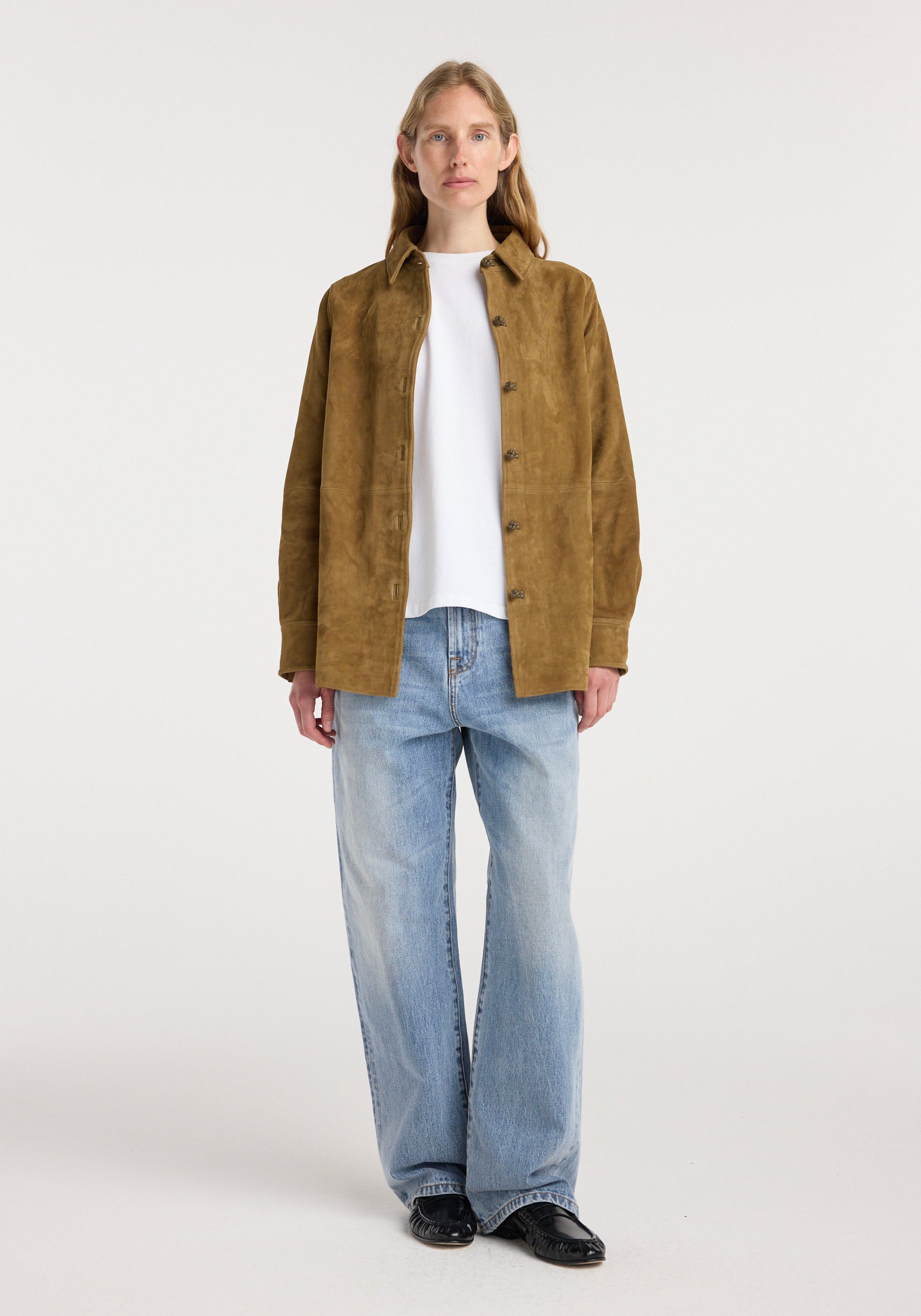 A woman wearing a brown suede pankou overshirt | sage over a white t-shirt paired with light blue wide-leg jeans and black shoes, standing against a plain background.