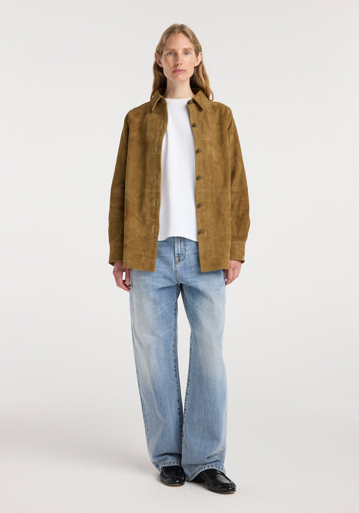A woman wearing a brown suede pankou overshirt | sage over a white t-shirt paired with light blue wide-leg jeans and black shoes, standing against a plain background.