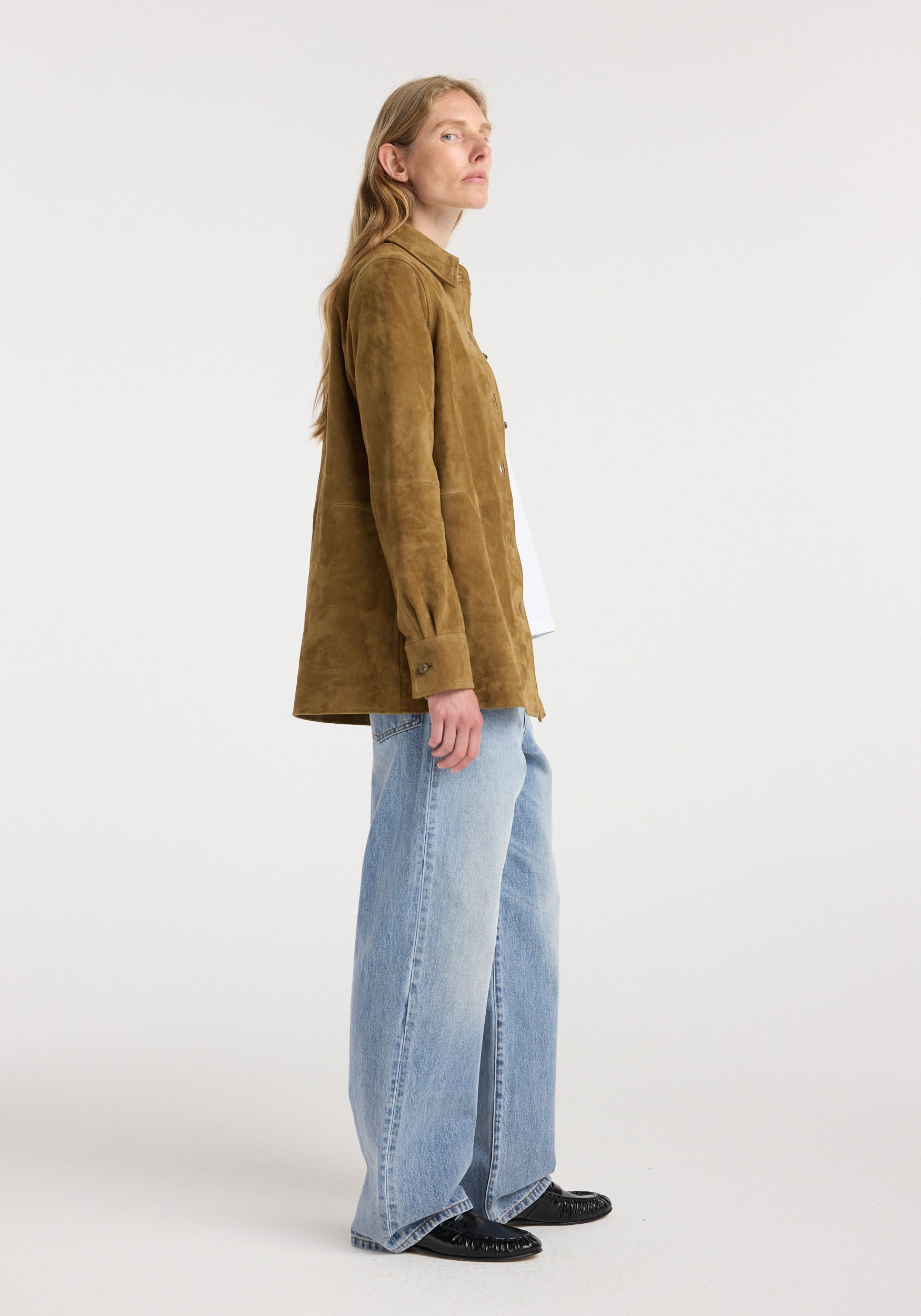 Side view of a woman wearing a brown suede pankou overshirt | sage with long sleeves, paired with light blue wide-leg jeans and black shoes against a plain light background.