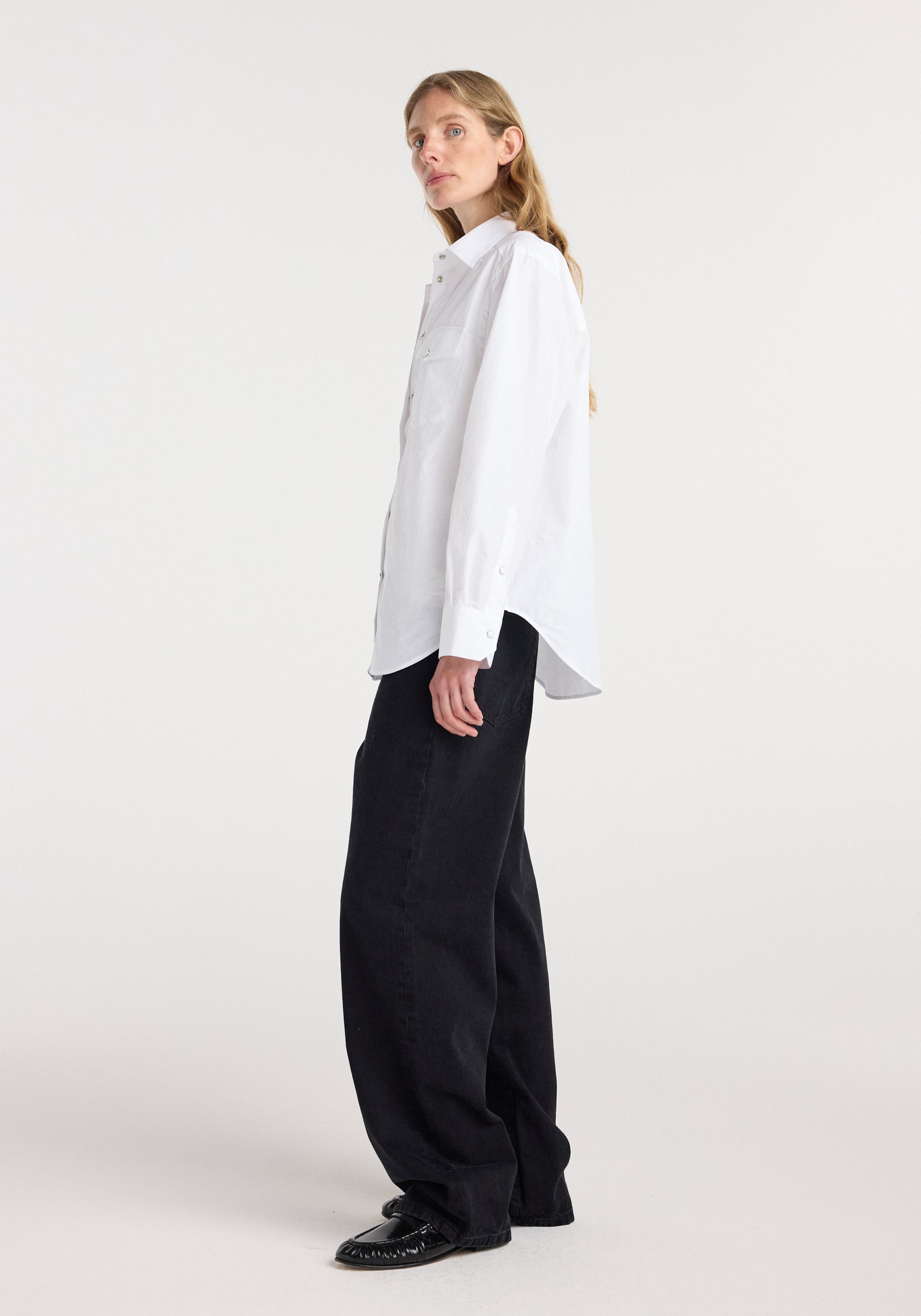 Side view of a woman wearing a loose-fitting white Western shirt | optic white paired with wide black pants and black shoes against a plain background.