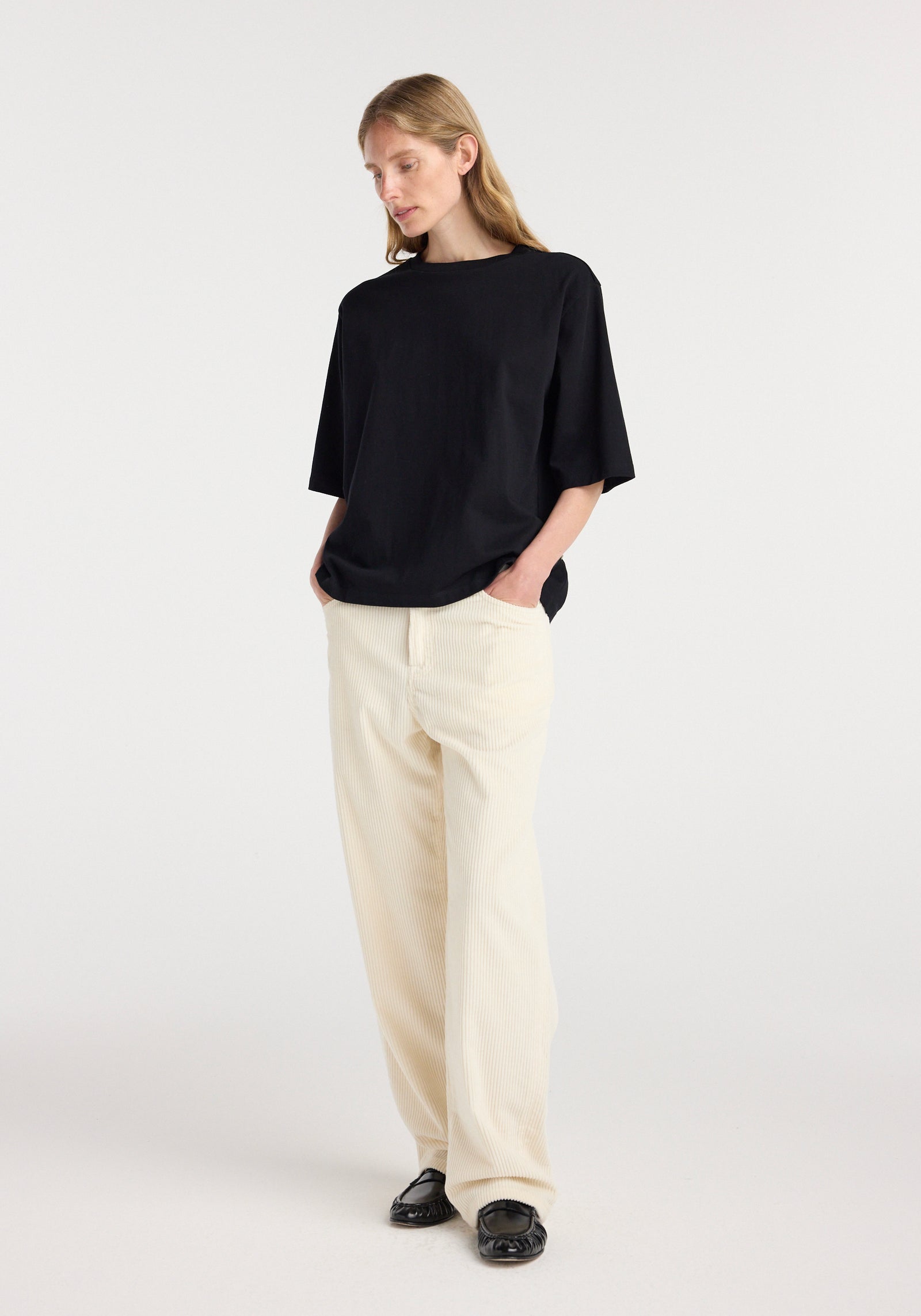Woman wearing an oversized cotton t-shirt | black with relaxed fit and hands in pockets, paired with light-colored wide-leg pants and black loafers, standing against a plain background.