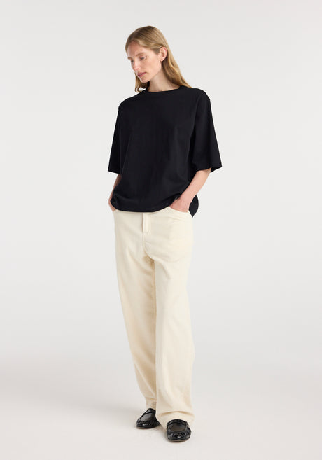 Woman wearing an oversized cotton t-shirt | black with relaxed fit and hands in pockets, paired with light-colored wide-leg pants and black loafers, standing against a plain background.