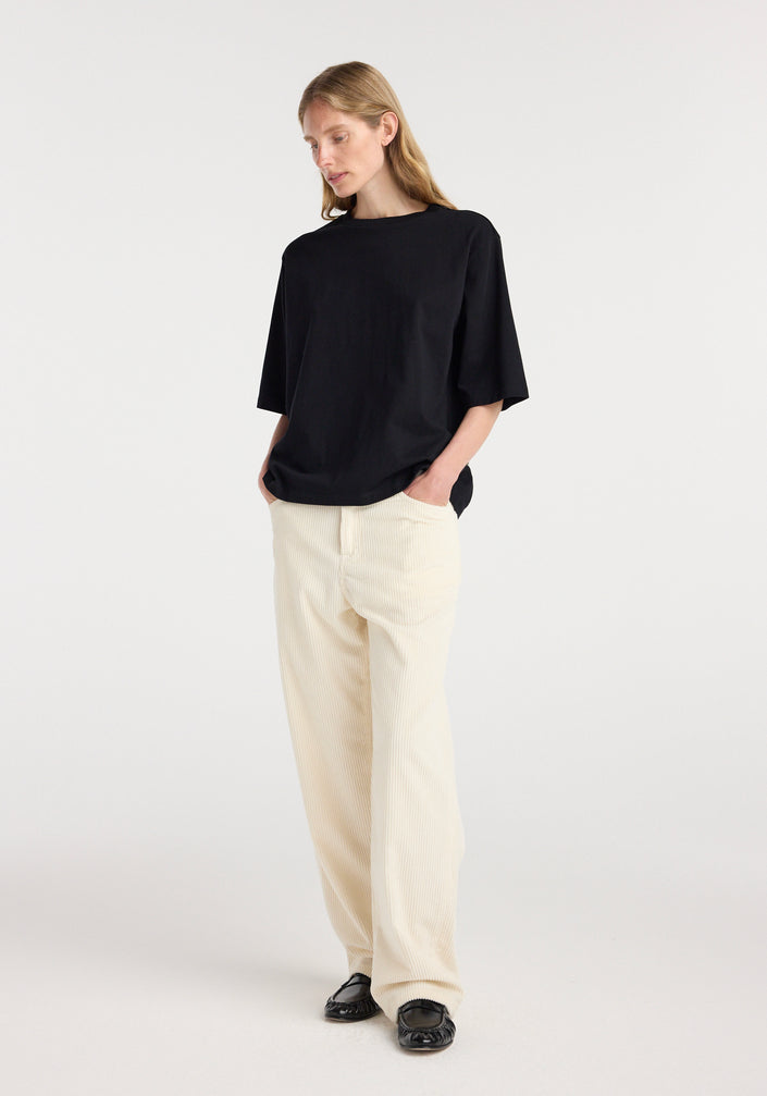 Woman wearing an oversized cotton t-shirt | black with relaxed fit and hands in pockets, paired with light-colored wide-leg pants and black loafers, standing against a plain background.