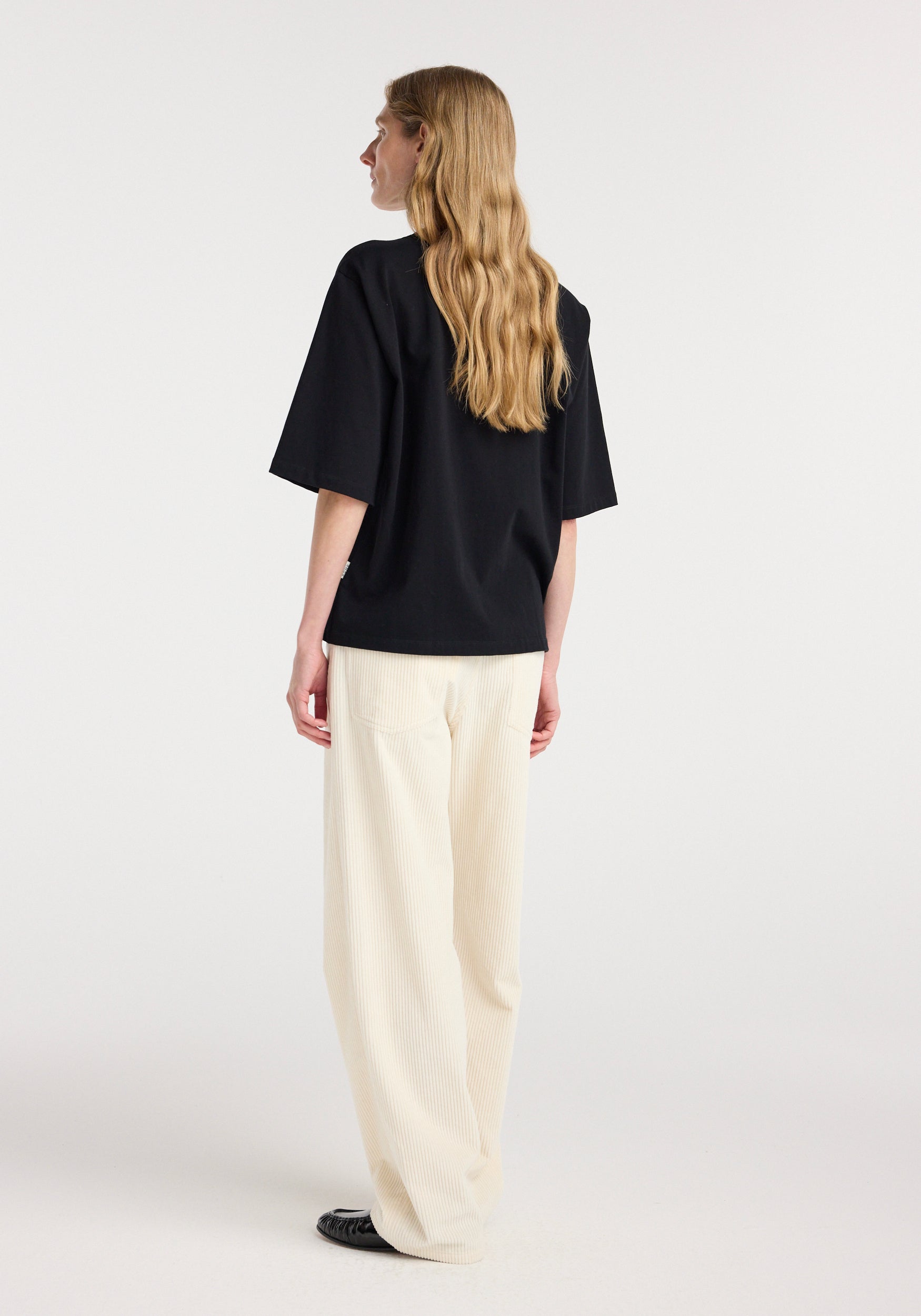 Woman with long blonde hair wearing an oversized cotton t-shirt | black, shown from the back, paired with loose white pants and black shoes against a plain background.