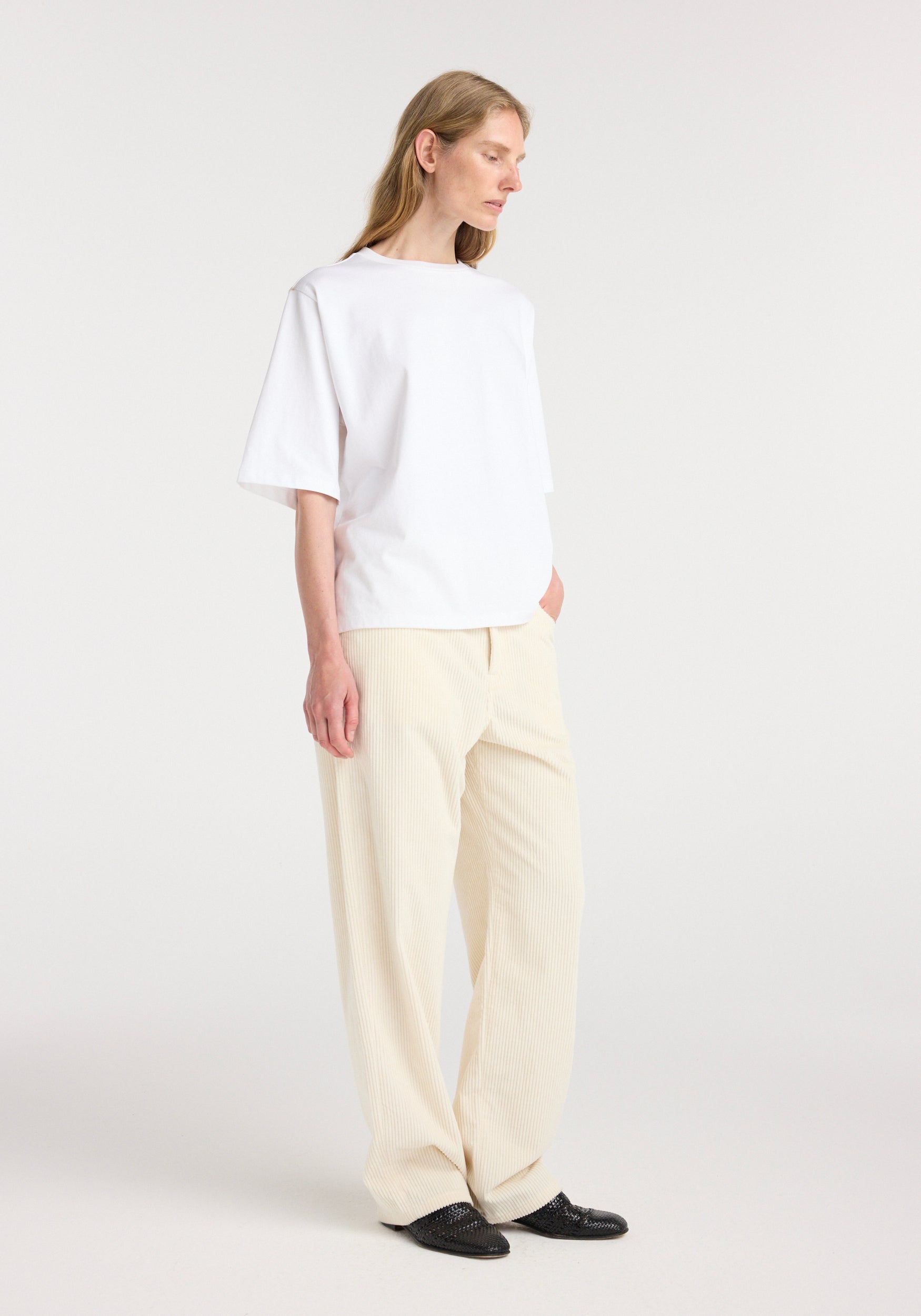 Woman wearing an oversized cotton t-shirt | optic white paired with light beige wide-leg pants and black pointed shoes standing against a plain white background.