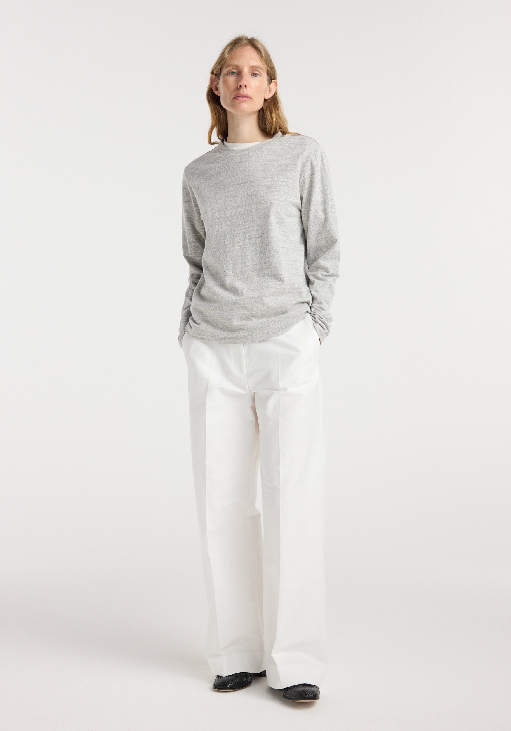 A model wearing a grey top paired with white wide-leg pants and black shoes, featuring the Japanese mouliné jersey long sleeve | grey mouliné in a simple, clean studio setting.