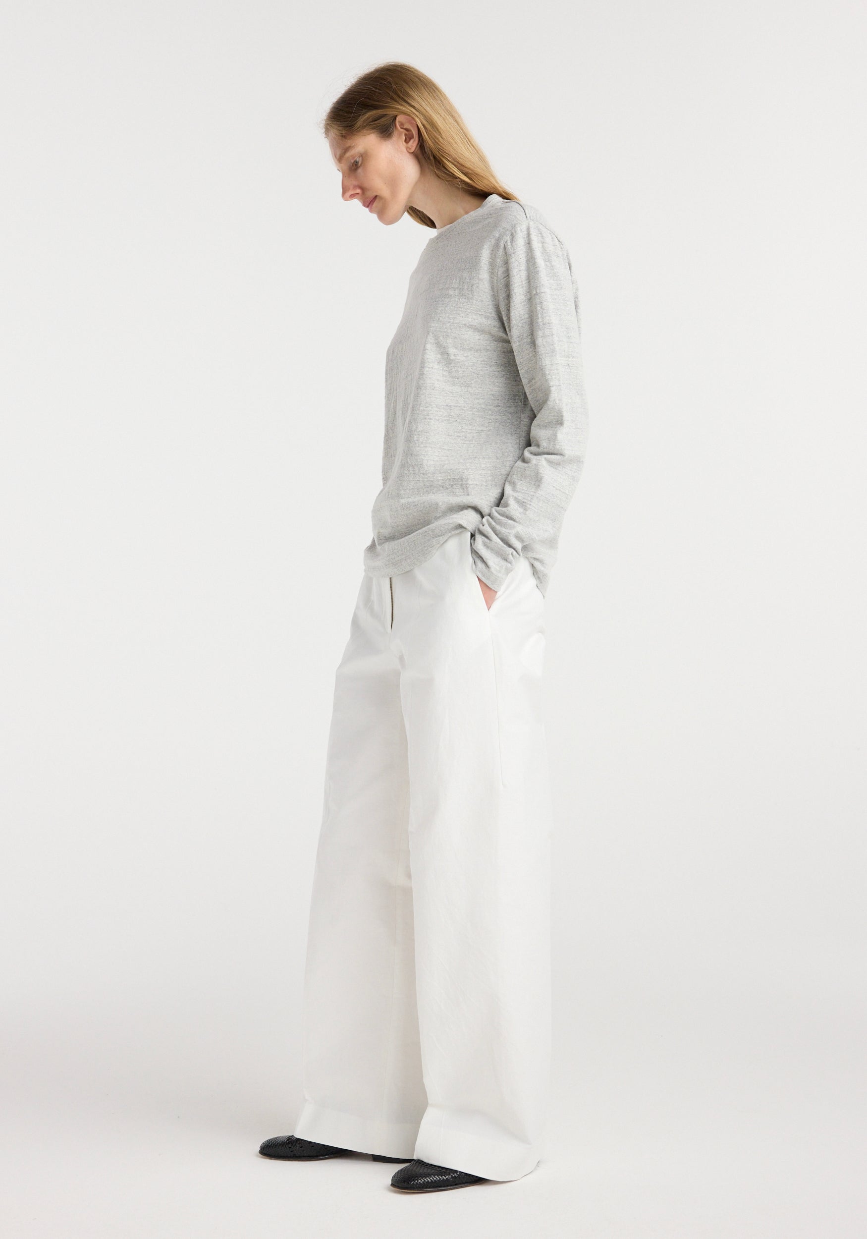Woman wearing a grey long sleeve top and wide white pants, standing with hands in pockets looking downward, styled in a minimalist setting, wearing Japanese mouliné jersey long sleeve | grey mouliné.