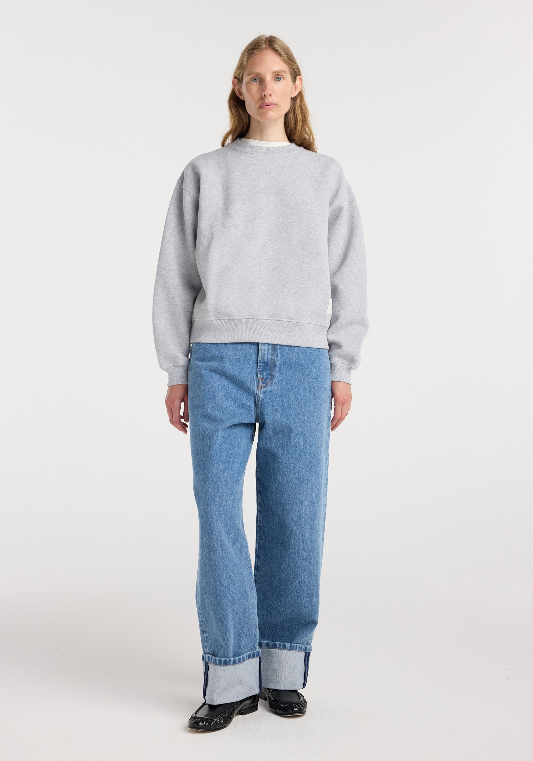 A woman wearing a light mid grey melange sweatshirt paired with high-waisted cuffed blue jeans and black shoes, standing against a plain white background in a studio.