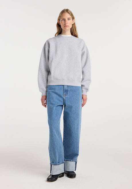 A woman wearing a light mid grey melange sweatshirt paired with high-waisted cuffed blue jeans and black shoes, standing against a plain white background in a studio.