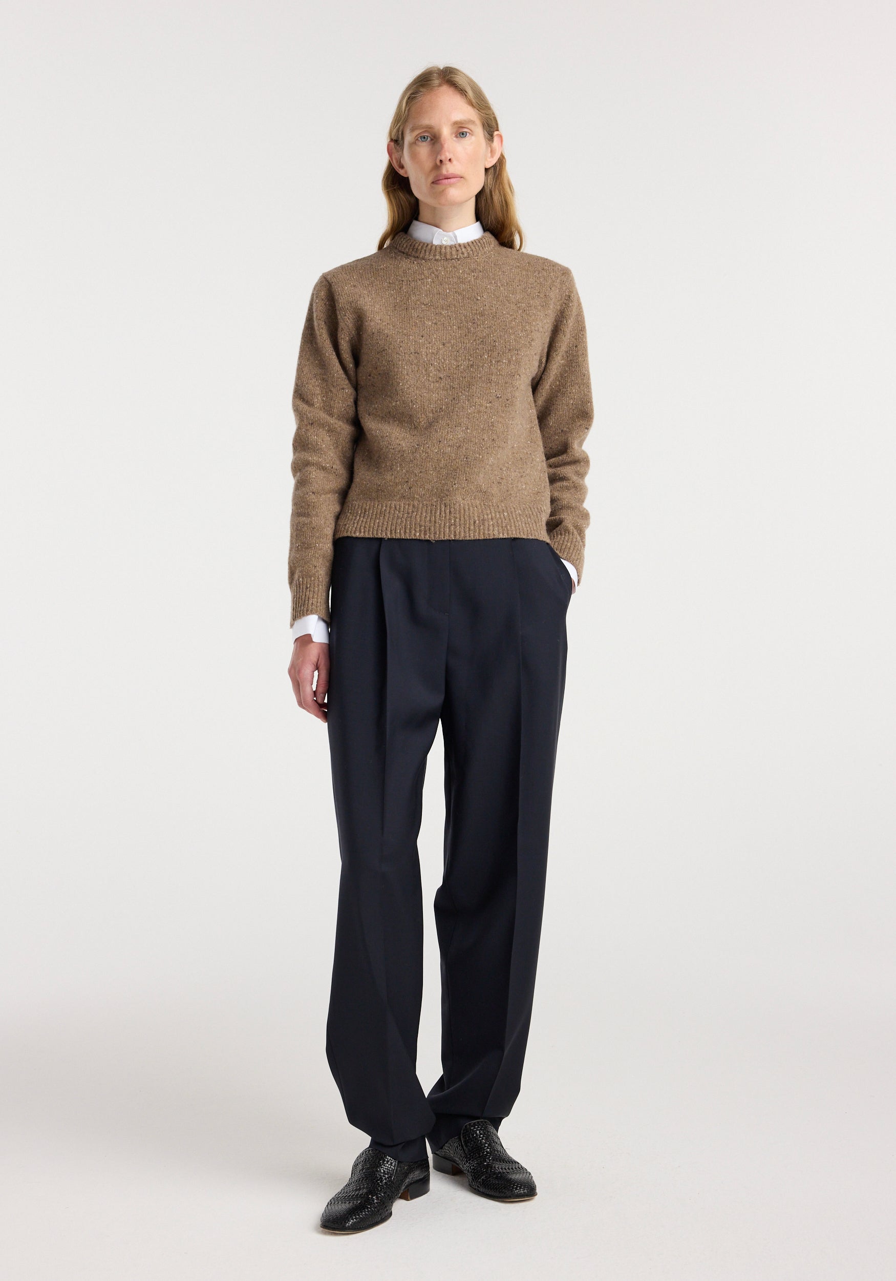 Woman wearing a fitted mouliné crewneck | mouliné taupe melange sweater with black pleated trousers and black shoes, standing against a plain white background.