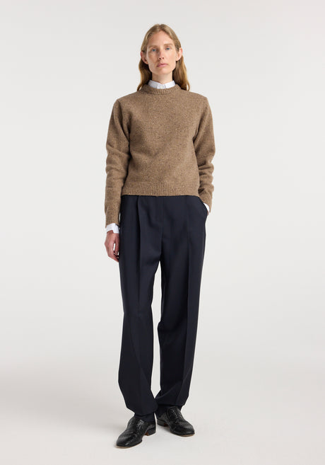 Woman wearing a fitted mouliné crewneck | mouliné taupe melange sweater with black pleated trousers and black shoes, standing against a plain white background.