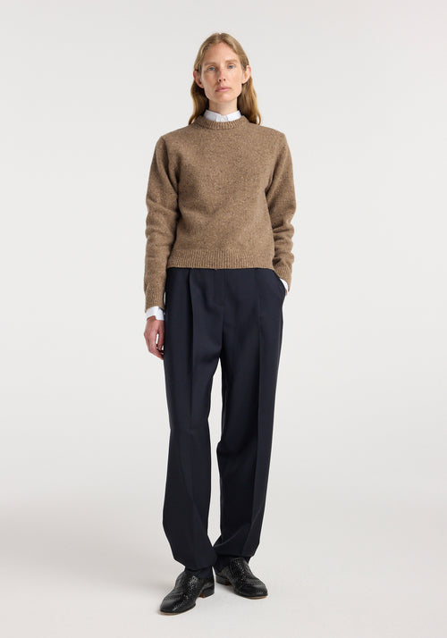 Woman wearing a fitted mouliné crewneck | mouliné taupe melange sweater with black pleated trousers and black shoes, standing against a plain white background.