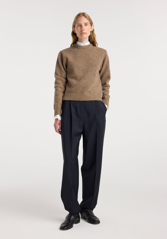 Woman wearing a fitted mouliné crewneck | mouliné taupe melange sweater with black pleated trousers and black shoes, standing against a plain white background.