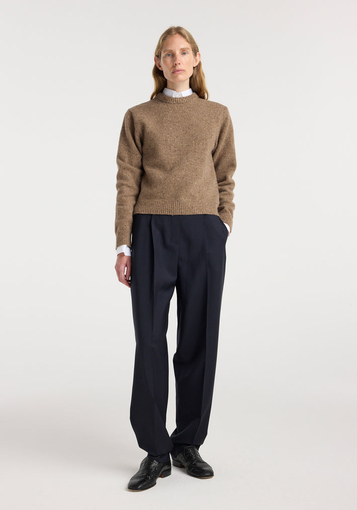 Woman wearing a fitted mouliné crewneck | mouliné taupe melange sweater with black pleated trousers and black shoes, standing against a plain white background.