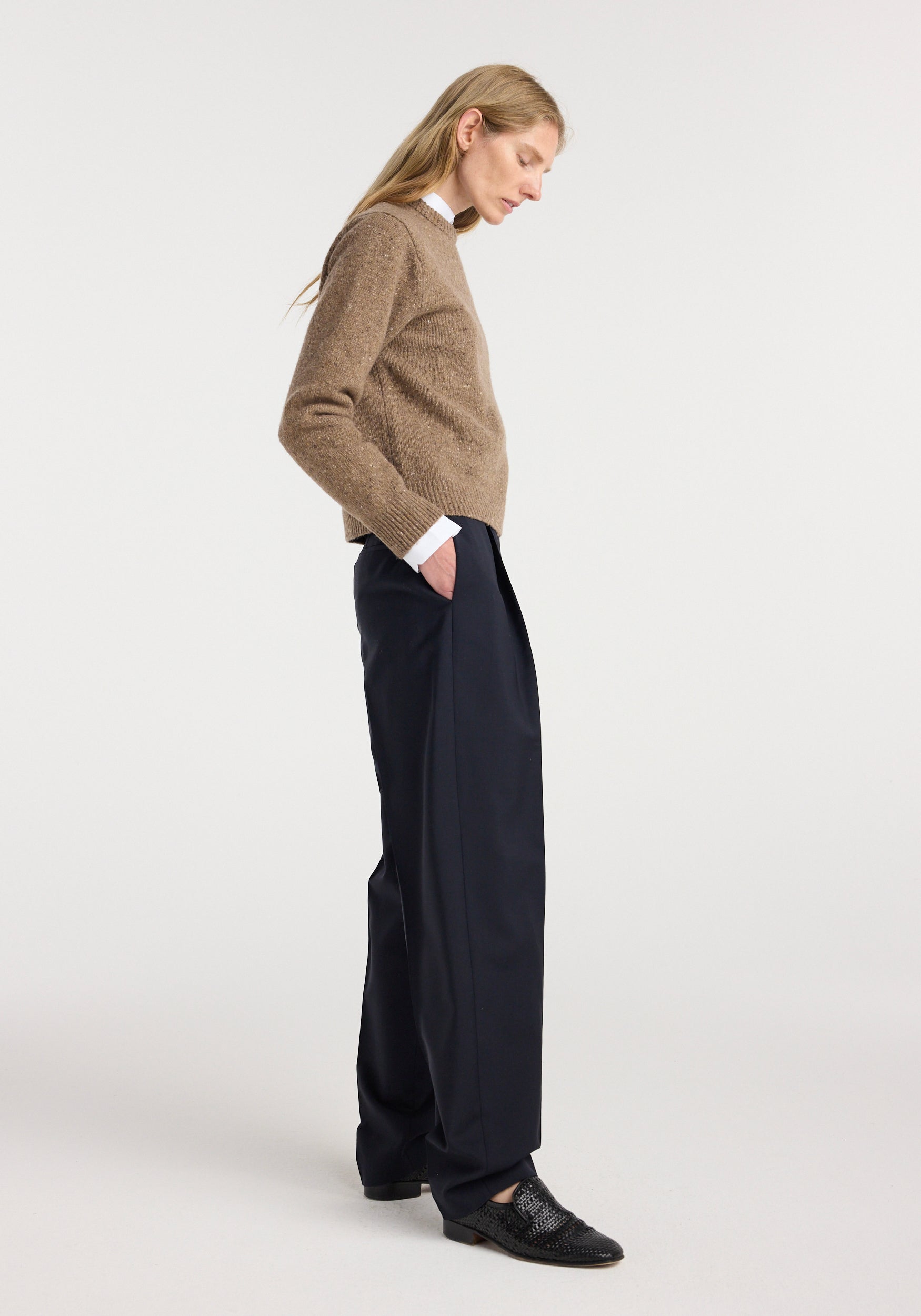Woman standing in profile wearing a fitted mouliné crewneck | mouliné taupe melange sweater paired with wide-leg black trousers and black shoes against a plain white background.