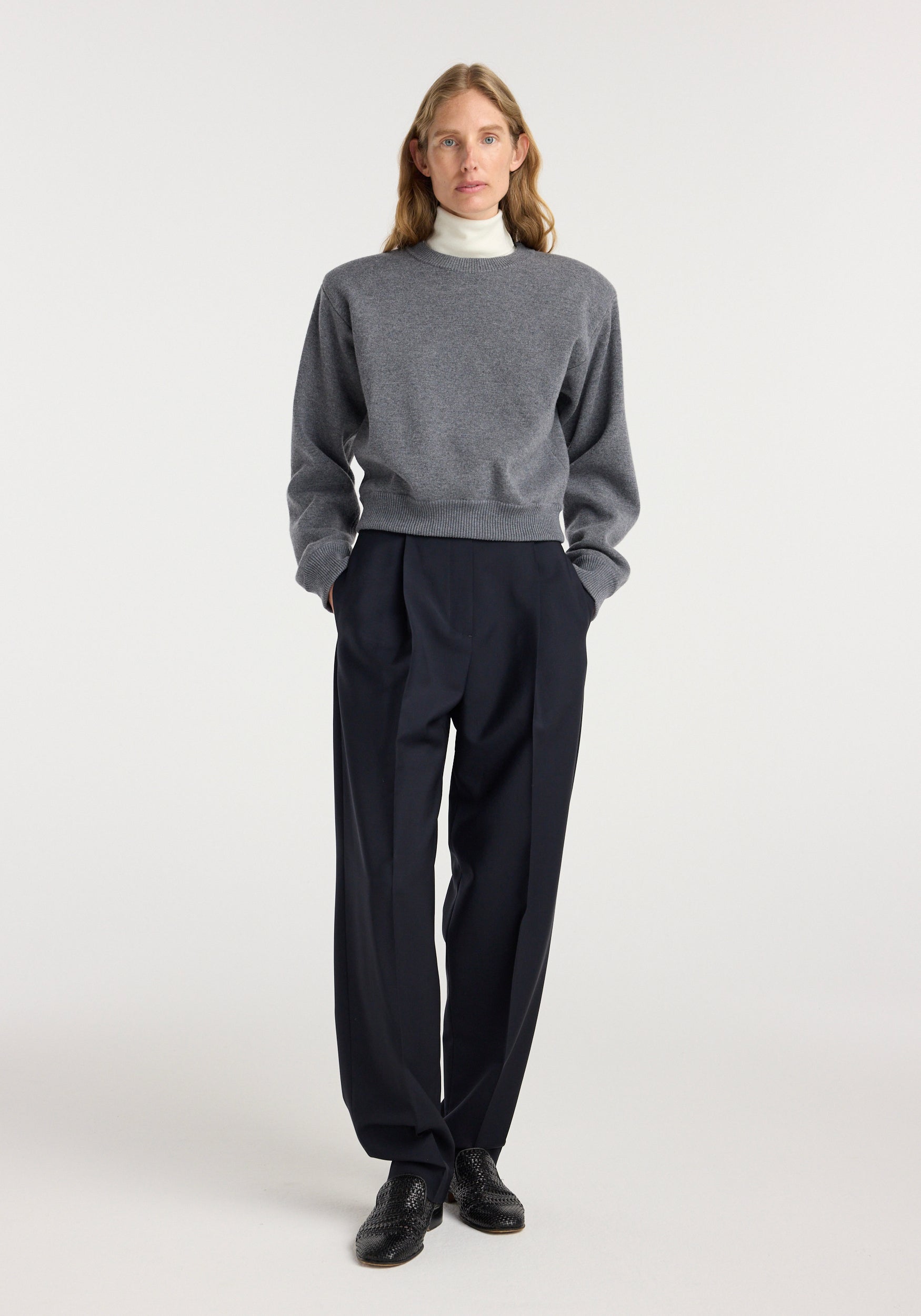 Woman wearing a mid grey melange Statement shoulder crewneck sweater over a white turtleneck, paired with black tailored wide-leg trousers and black shoes, standing against a plain background.