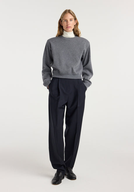 Woman wearing a mid grey melange Statement shoulder crewneck sweater over a white turtleneck, paired with black tailored wide-leg trousers and black shoes, standing against a plain background.