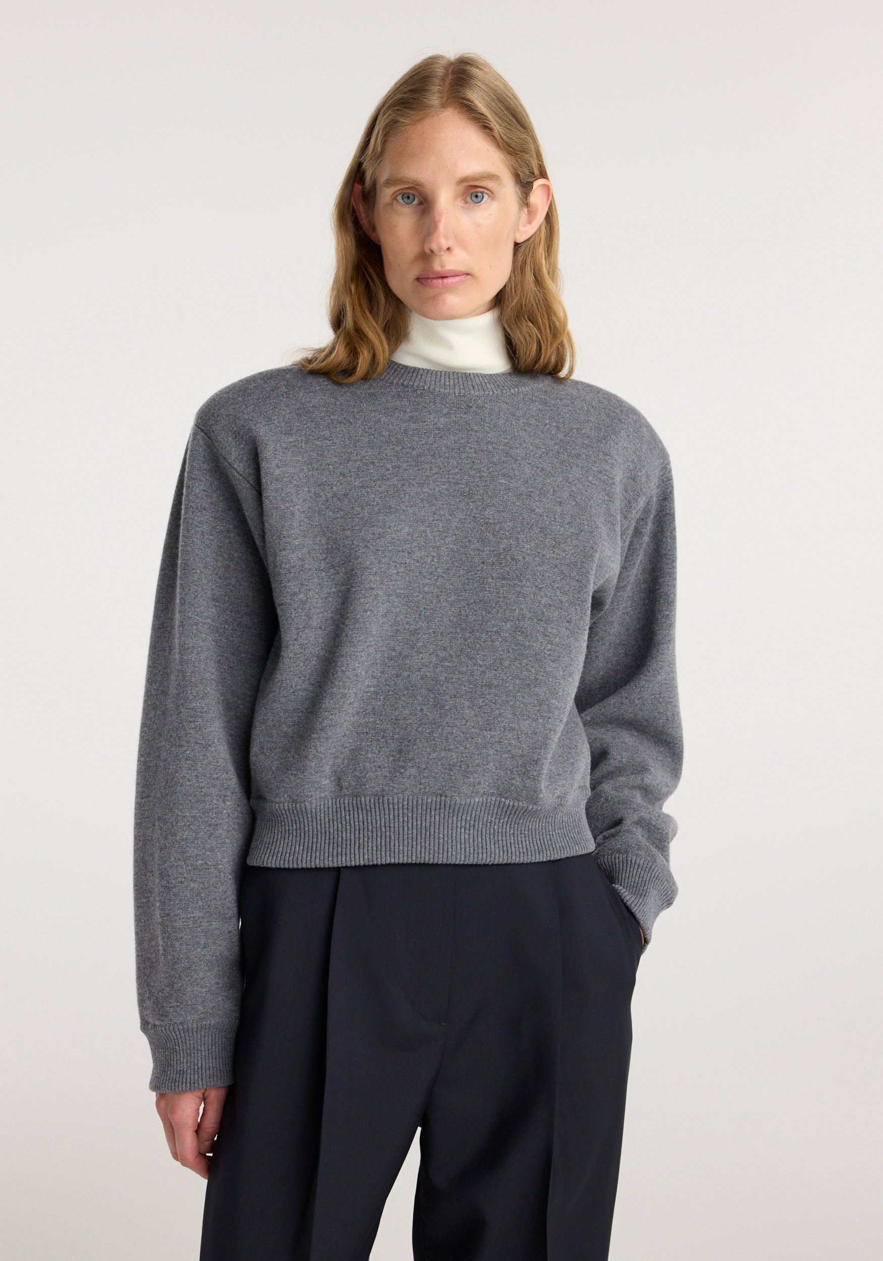 Woman wearing a mid grey melange sweater with a statement shoulder design over a white turtleneck, paired with black trousers, showcasing the Statement shoulder crewneck | mid grey melange.