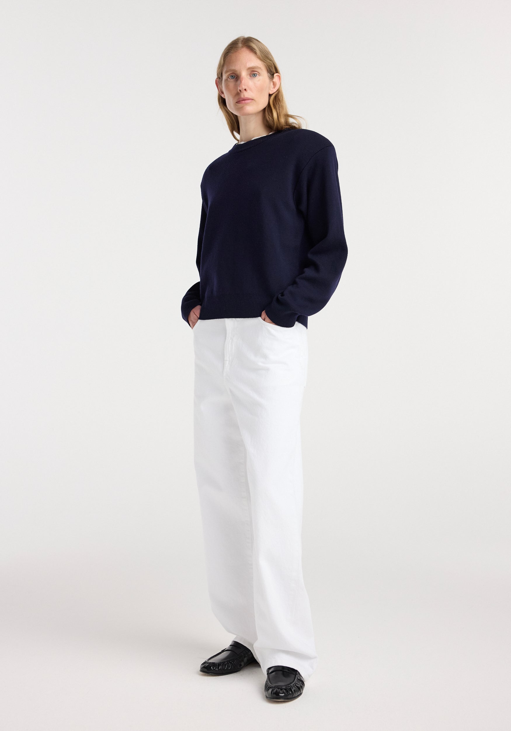 Woman wearing a navy Statement shoulder crewneck | navy sweater paired with loose white pants and black shoes, standing against a plain light background.