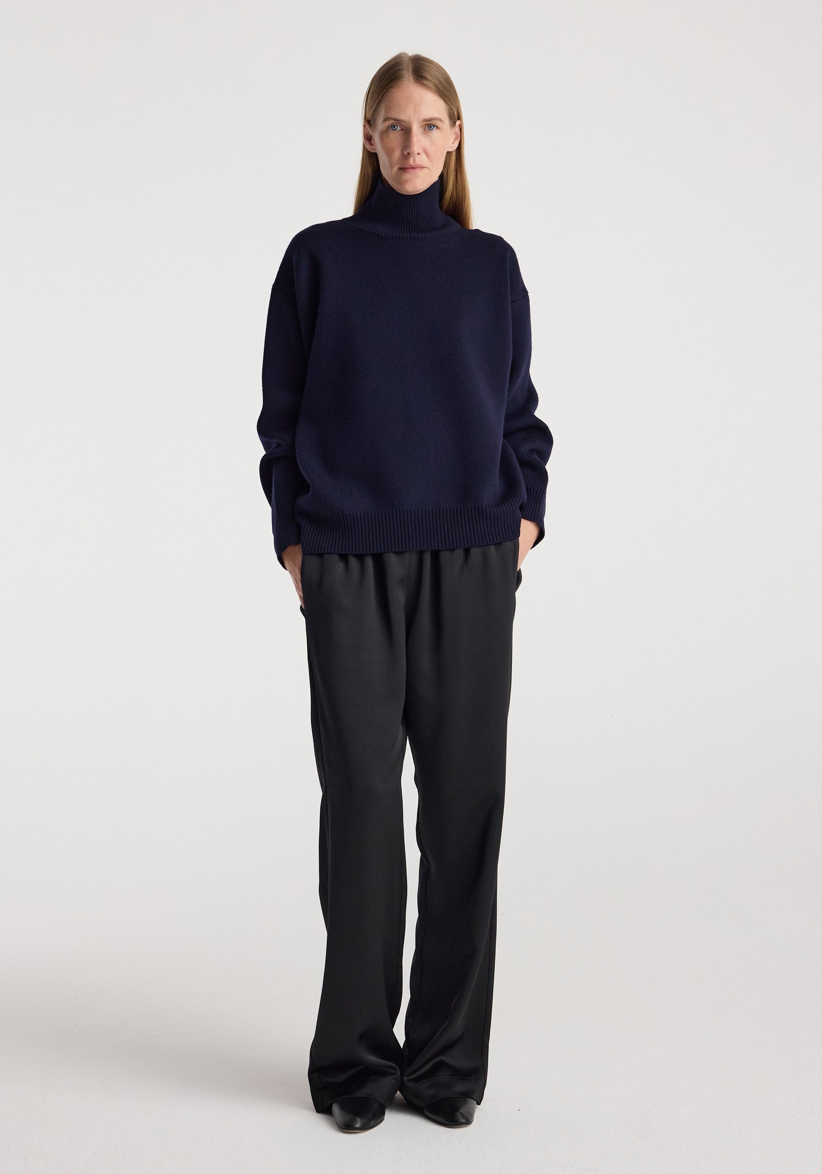 Woman wearing a navy sculptural compact turtleneck with long sleeves paired with loose black pants, standing against a plain white background.