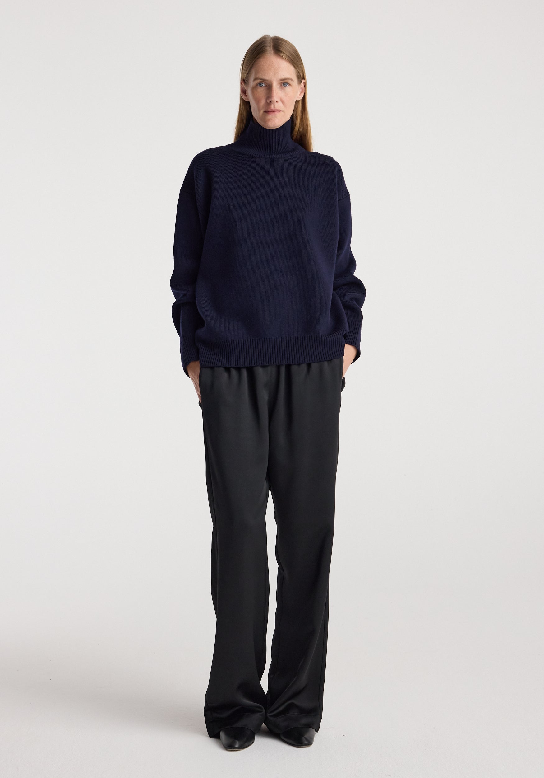 Woman wearing a navy sculptural compact turtleneck with long sleeves paired with loose black pants, standing against a plain white background.