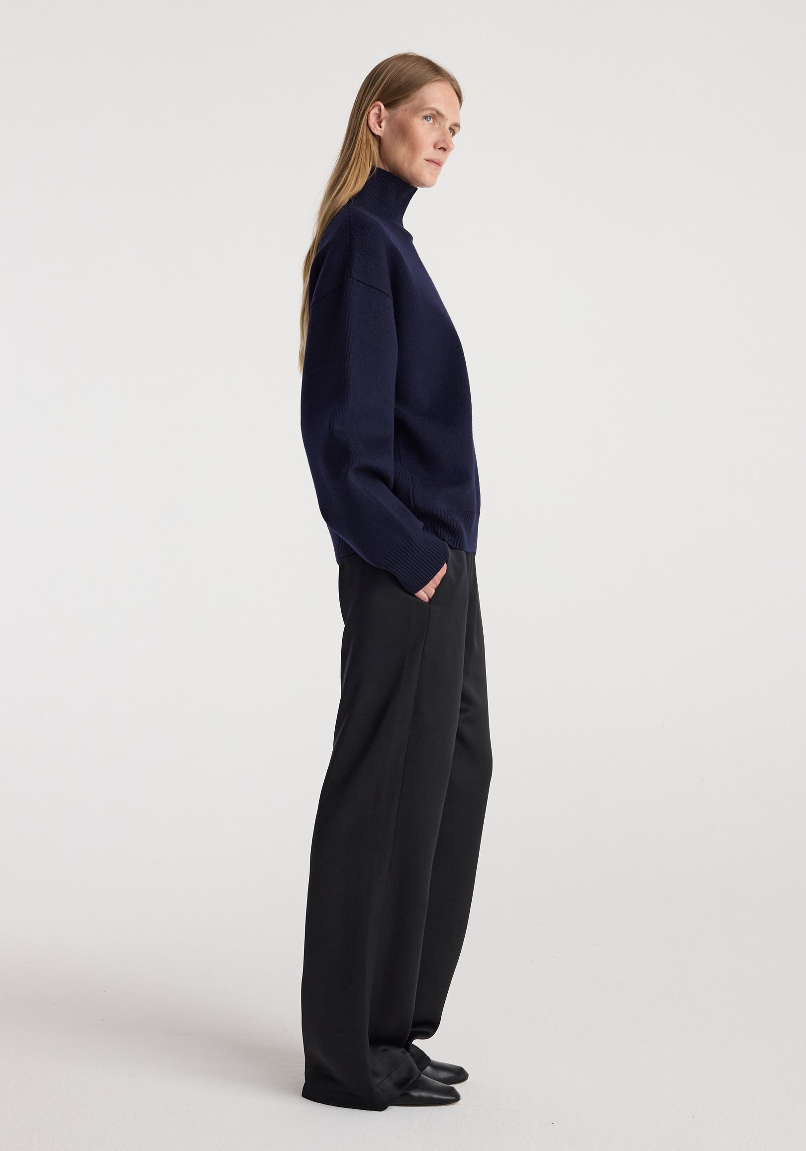 Side view of a woman wearing a navy sculptural compact turtleneck paired with loose black trousers and black shoes against a plain background.