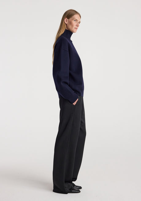 Woman wearing a navy sculptural compact turtleneck with long sleeves paired with loose black pants, standing against a plain white background.