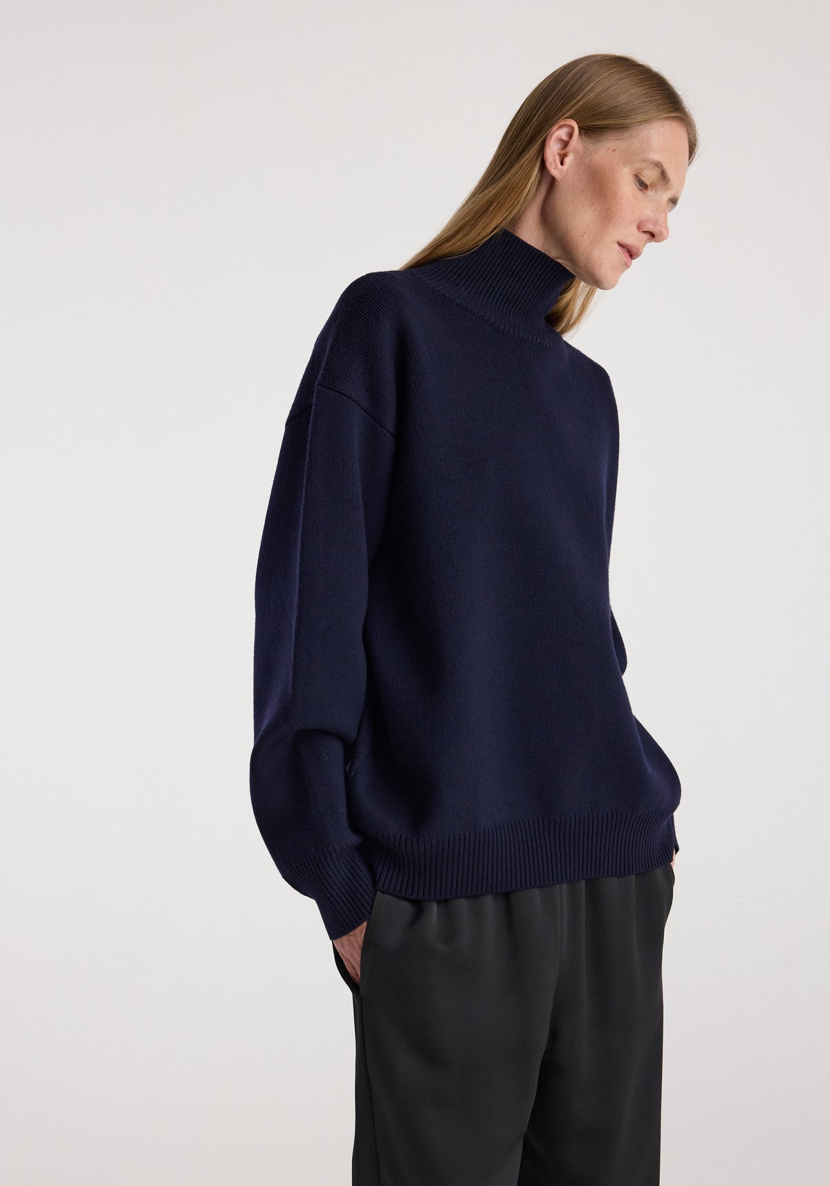 A woman wearing a navy sculptural compact turtleneck with dropped shoulders and long sleeves, paired with loose black pants, standing against a plain background.