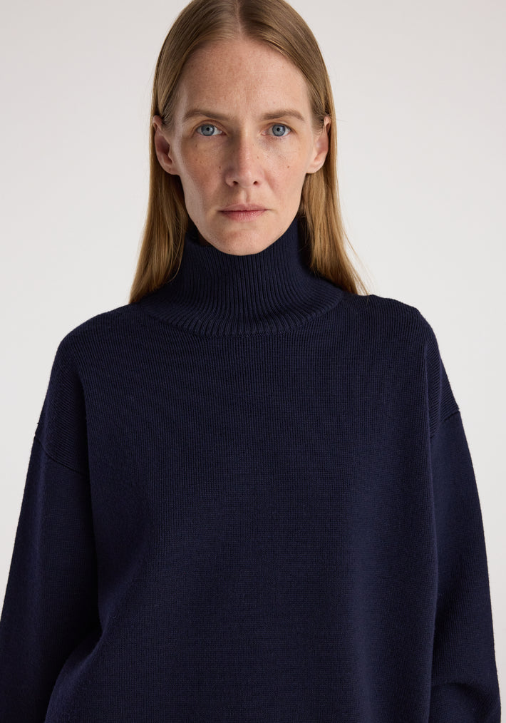 Front view of a woman wearing a navy blue Sculptural compact turtleneck | navy sweater with a ribbed high neck and relaxed fit against a plain light background.